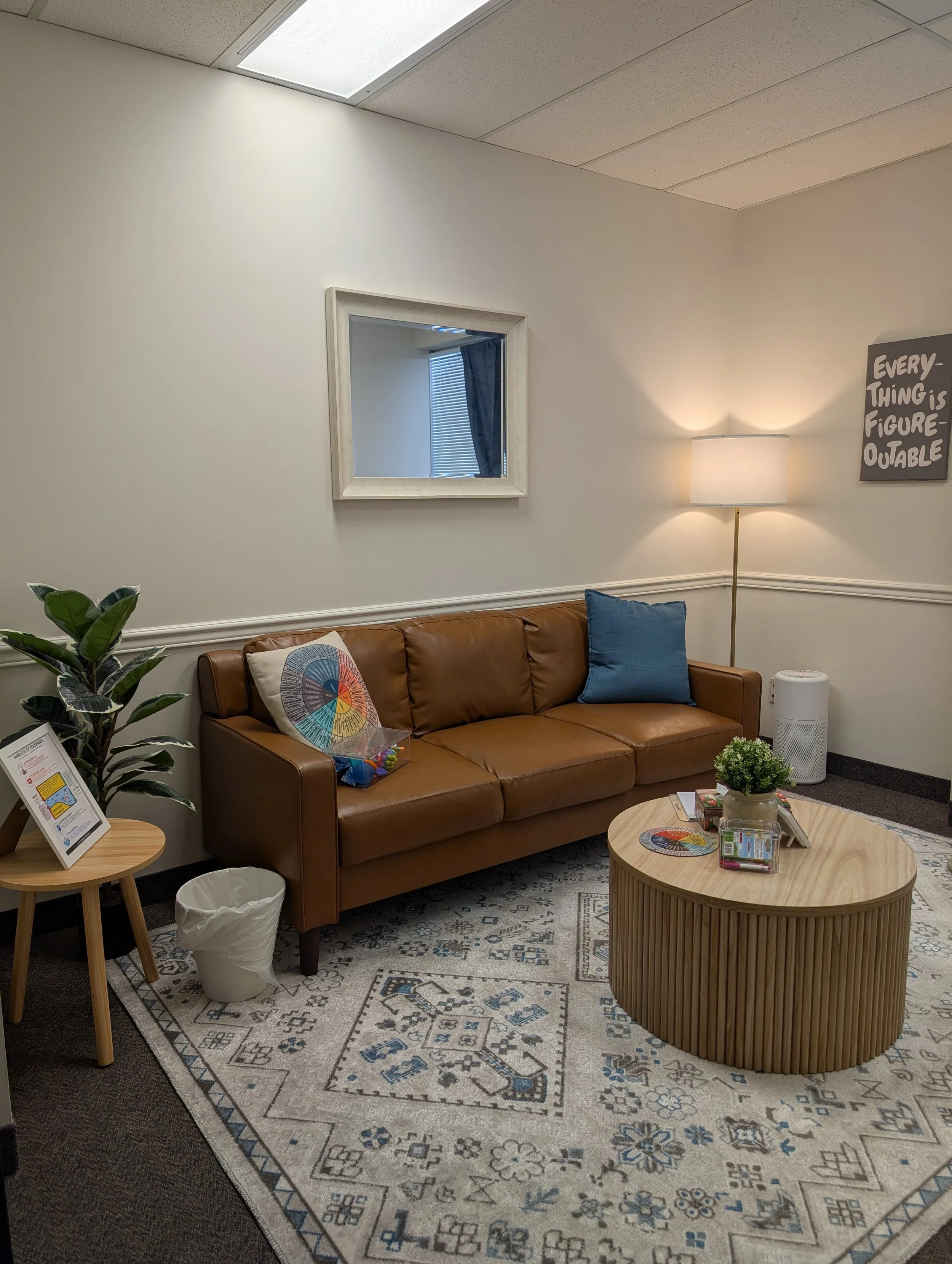 A cozy waiting room with a brown leather sofa, a coffee table with magazines, and a decorative rug. A tall floor lamp and a plant are also present, along with a mirror on the wall and a framed quote that says "Every-thing is figure-outable."