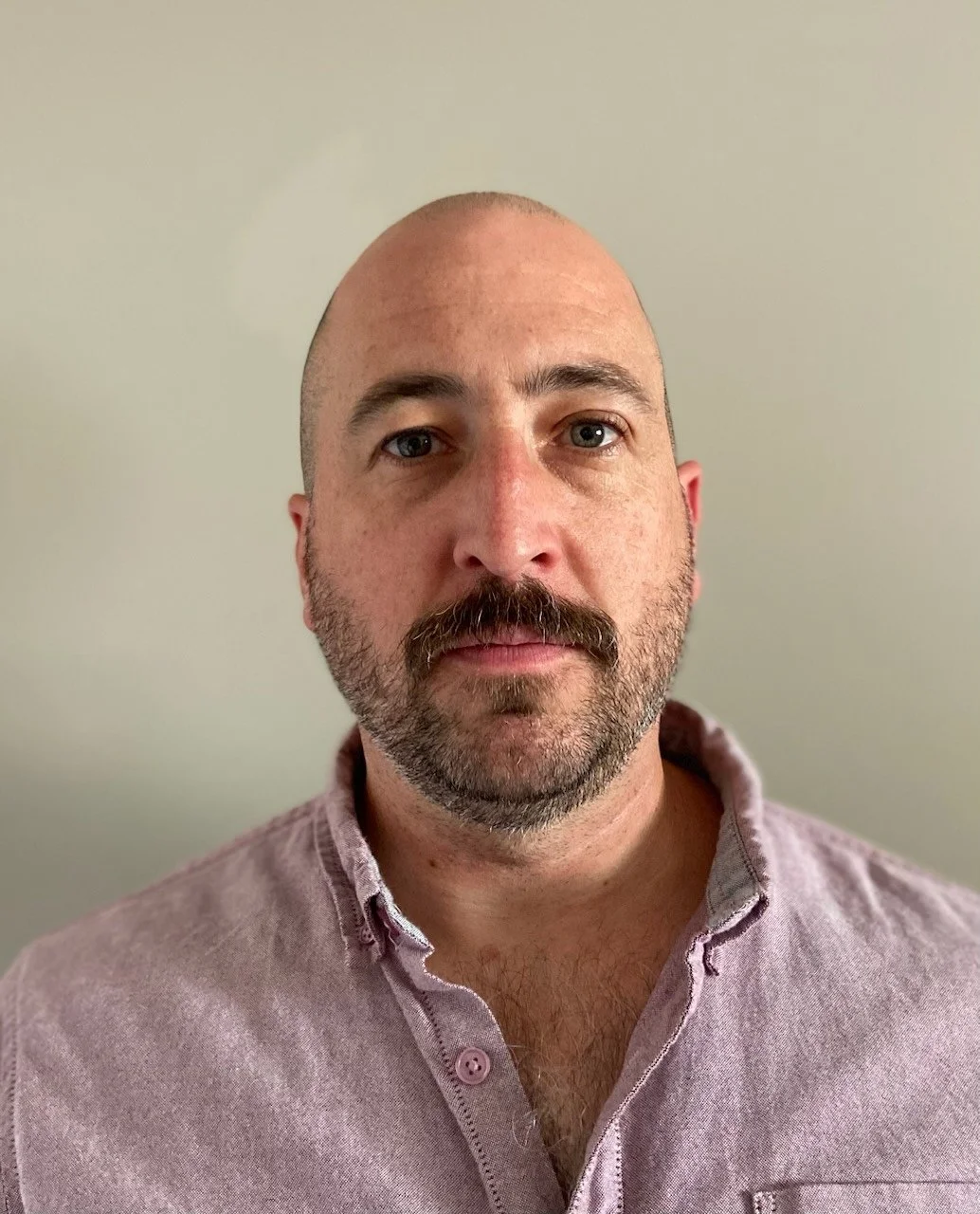 Headshot of a bald man with a beard and mustache, wearing a light pink collared shirt against a plain wall background.