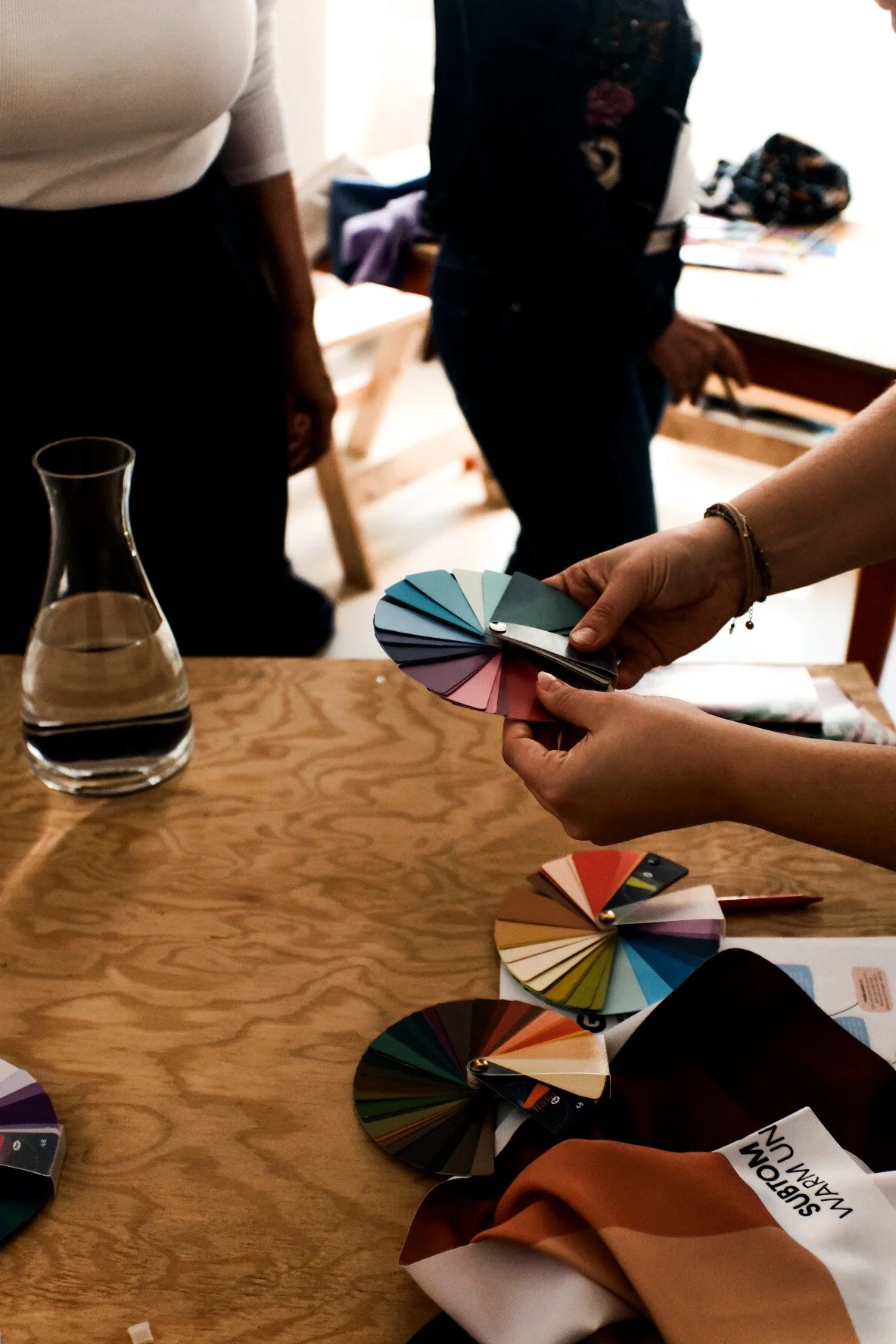 Hände, die Farbtafeln in verschiedenen Farben halten, auf einem Holztisch mit weiteren Farbfächern bei einem Farbanalyse-Workshop von Studio Barb.