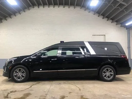 Black hearse parked inside a warehouse with a white brick wall in the background.