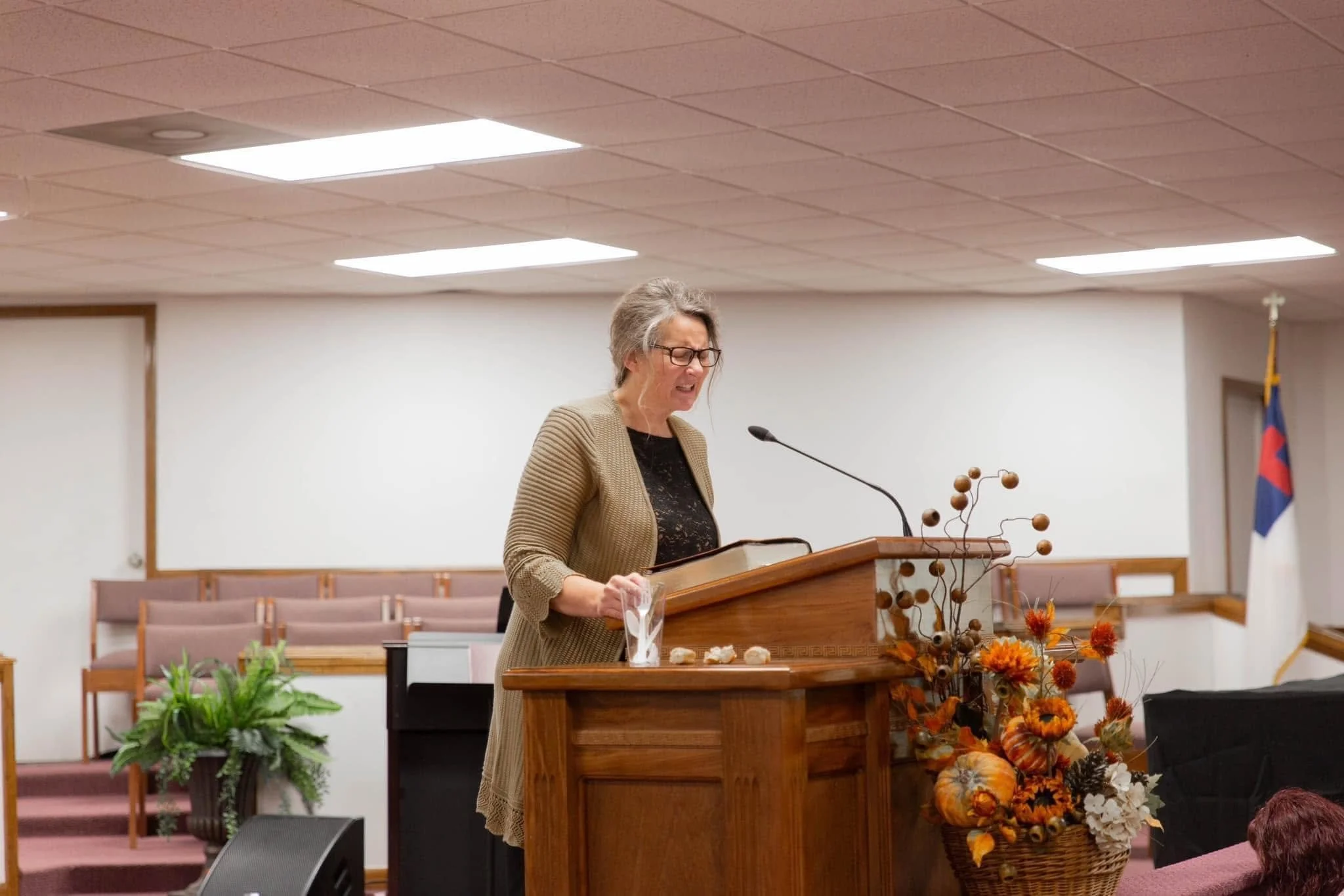 A woman with gray hair and glasses is speaking at a wooden podium decorated with autumn-colored flowers and dried branches in a church or conference room. She is wearing a beige blazer over a black top. There are empty chairs and plants in the backgr