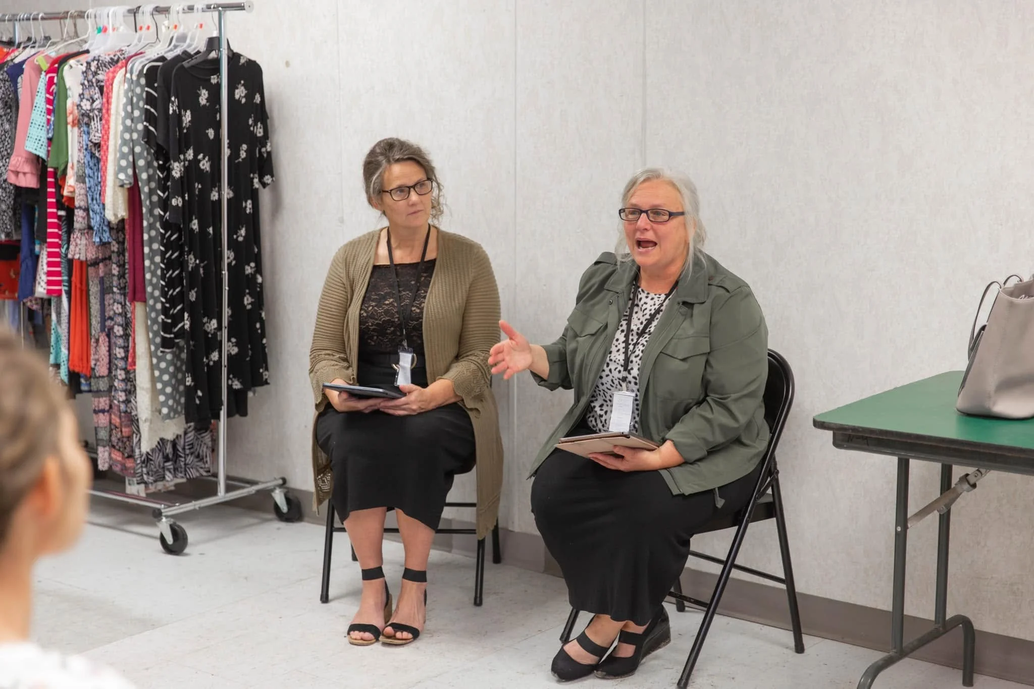 Two women sitting and talking; one holding a tablet, the other speaking with hand gestures. Clothing rack with colorful dresses in the background.
