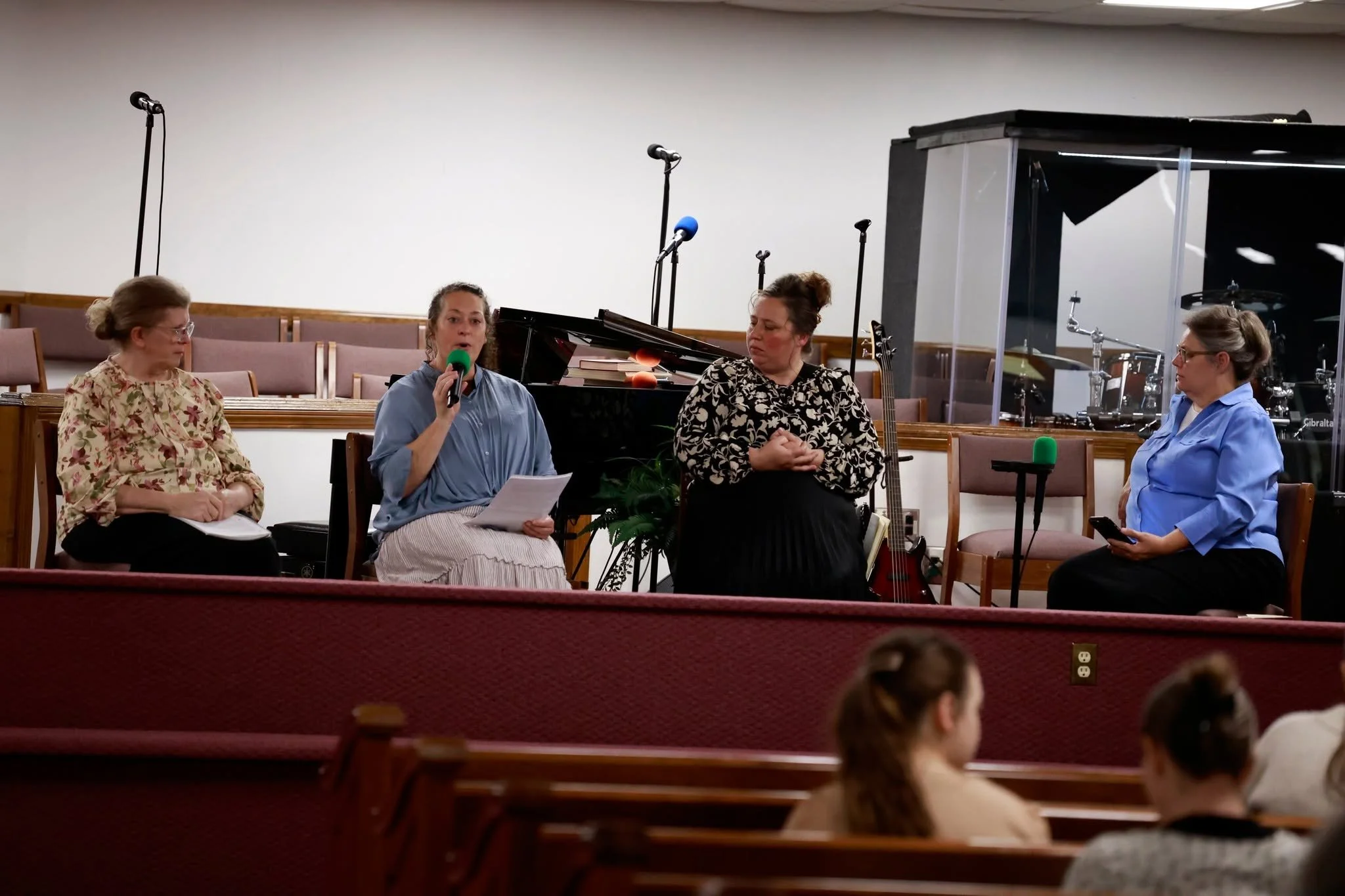 Four women sitting in a church, engaging in a discussion or interview, with a grand piano, microphones, books, and musical instruments in the background.