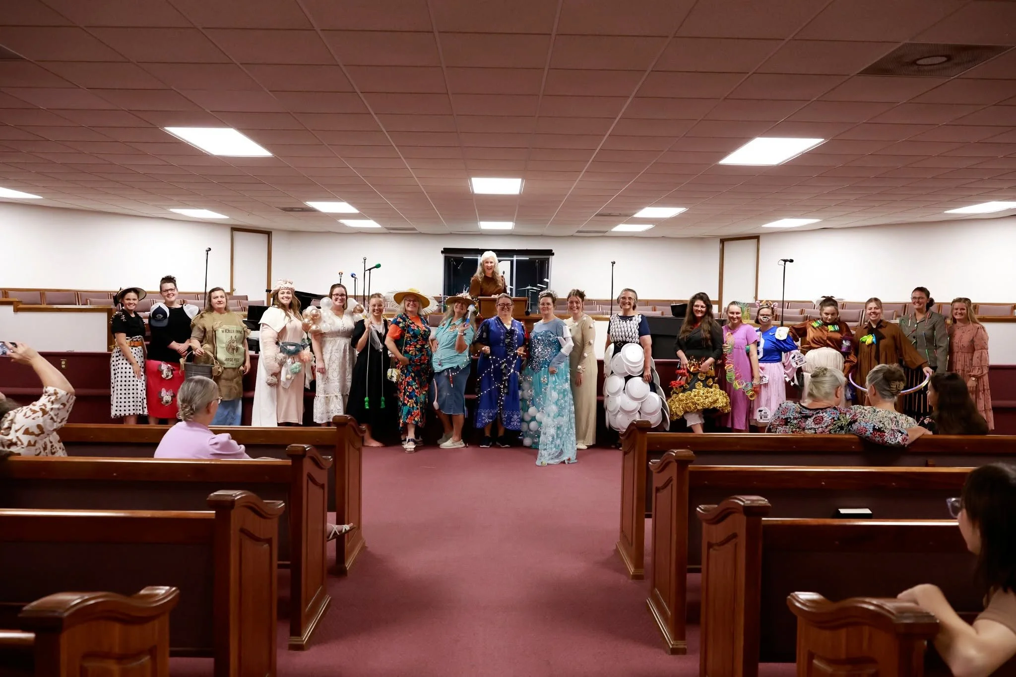Group of women dressed in costumes standing on a church stage, with some holding props, and a woman speaking at a podium in front of them, with a congregation seated in pews.