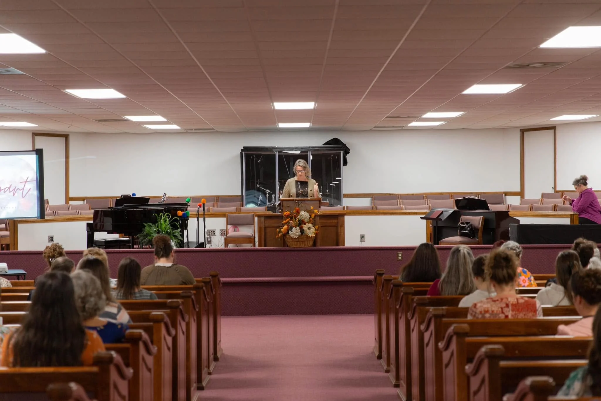 A woman stands at a lectern speaking in a church or meeting hall with a congregation seated in pews. The room has a wood-paneled ceiling with fluorescent lighting and a stage area with musical instruments and a large basket of flowers.
