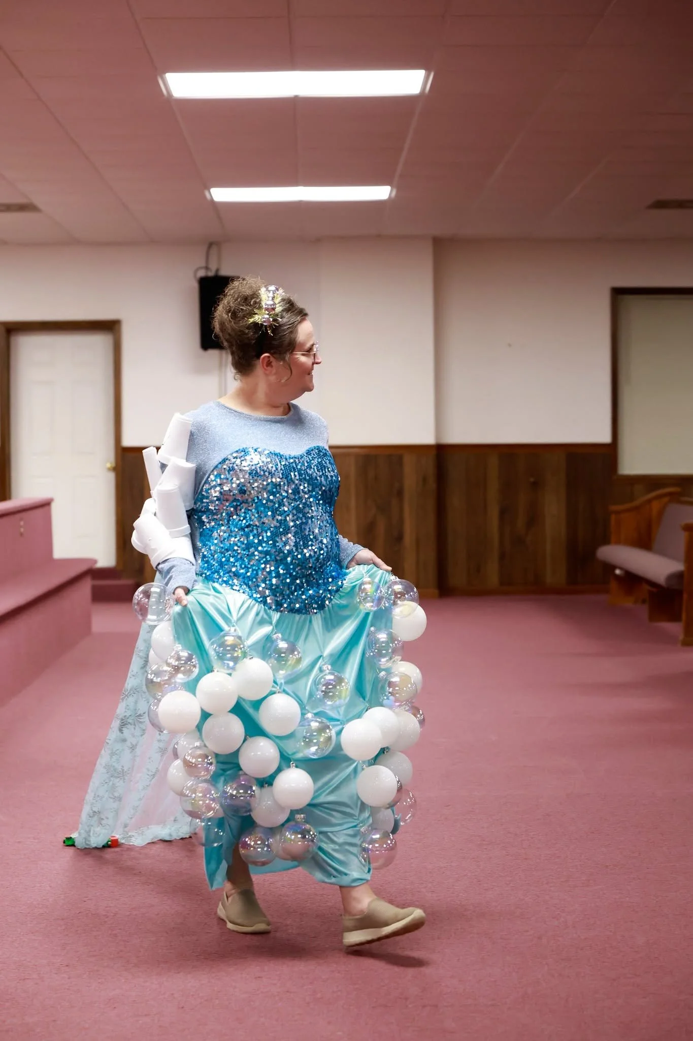 A woman dressed in a shiny, light blue dress decorated with clear and white balloons, walking across a room with pink carpet and wood-paneled walls, wearing beige shoes and glasses, with a floral headband in her curly hair.