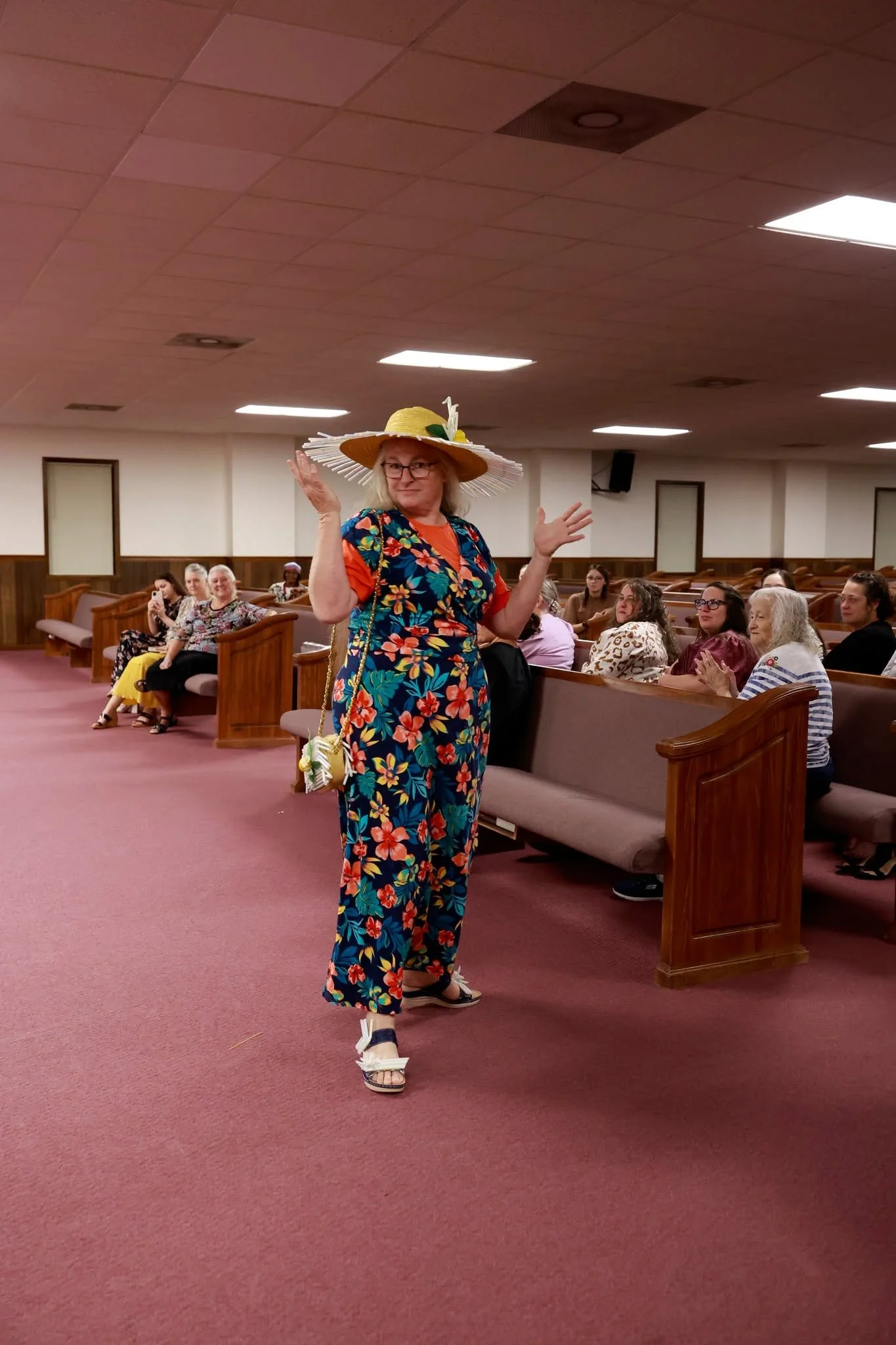 A woman wearing a large yellow sunhat decorated with white and green accents, a colorful floral jumpsuit, and sandals. She is standing in a church or auditorium, surrounded by seated people, some smiling and clapping.