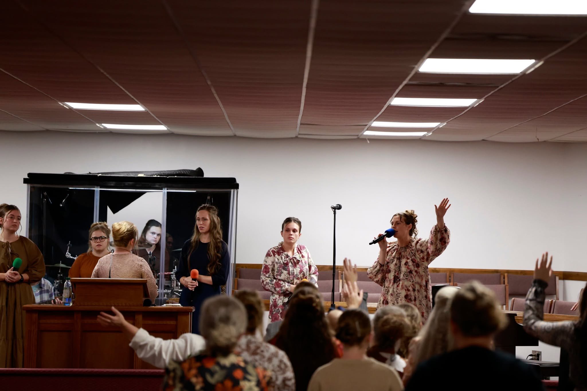 A group of women on stage, one is speaking into a microphone and raising her hand, while others hold microphones in a church or community setting, with audience members raising their hands.