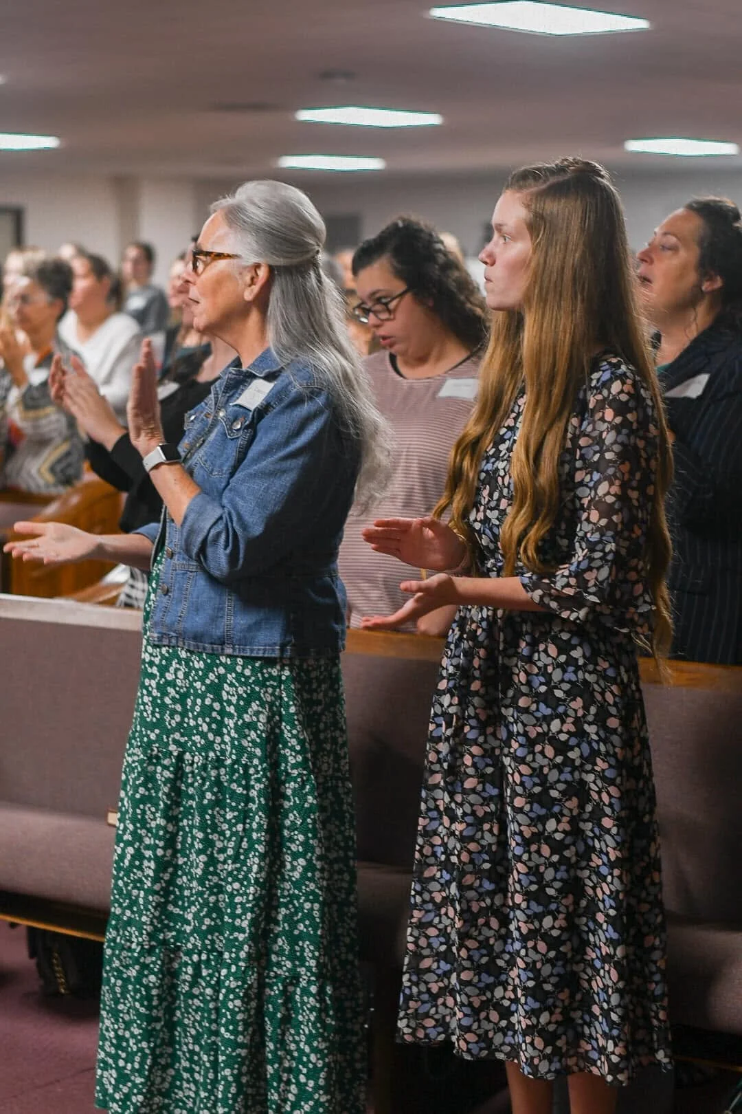 A diverse group of women standing in a church or auditorium, praying or worshipping with their hands raised, during a religious service.