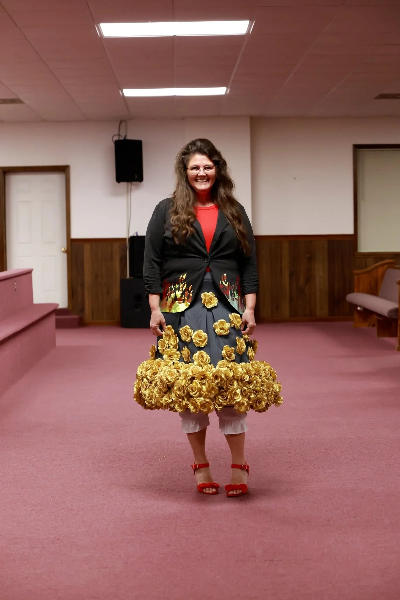 A woman standing in a room with pink carpet and wood-paneled walls, wearing a unique dress made of yellow roses, red shoes, and glasses, smiling at the camera.