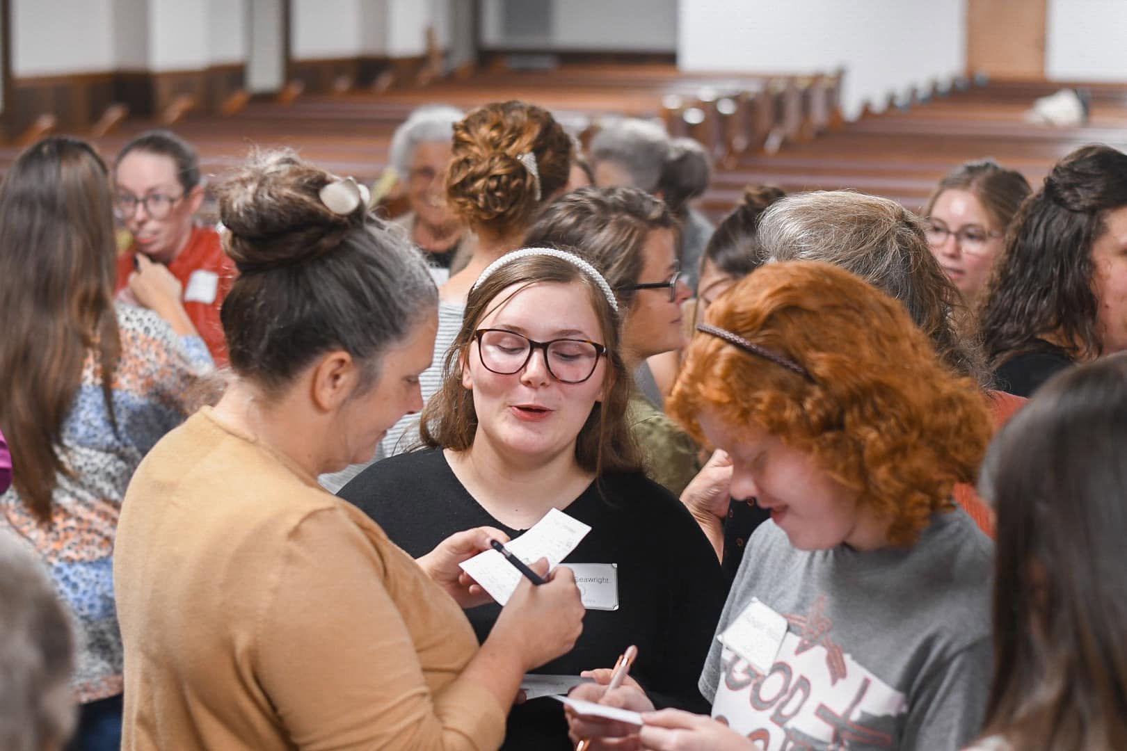 A group of women gathered together, some with glasses and various hairstyles, engaging in conversation and writing notes inside a venue with wooden pews.