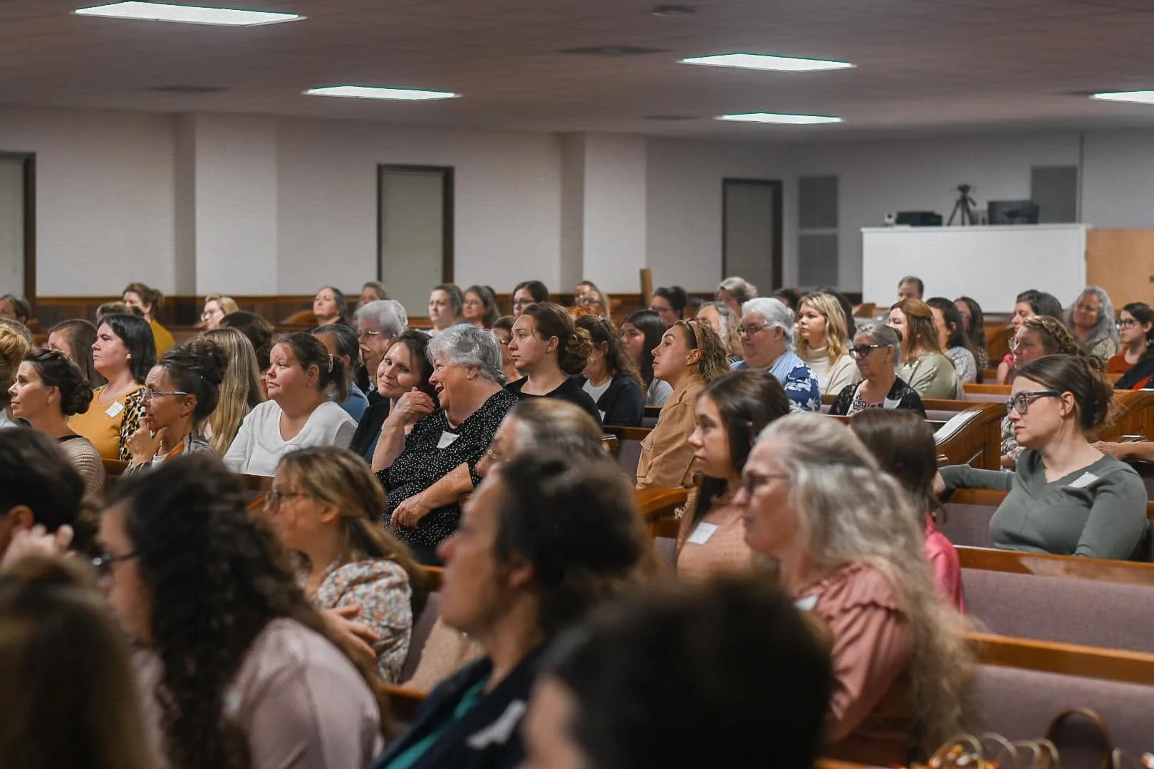 An audience of diverse women and men seated in rows of pews in a conference room, attentively listening to a speaker or presentation.