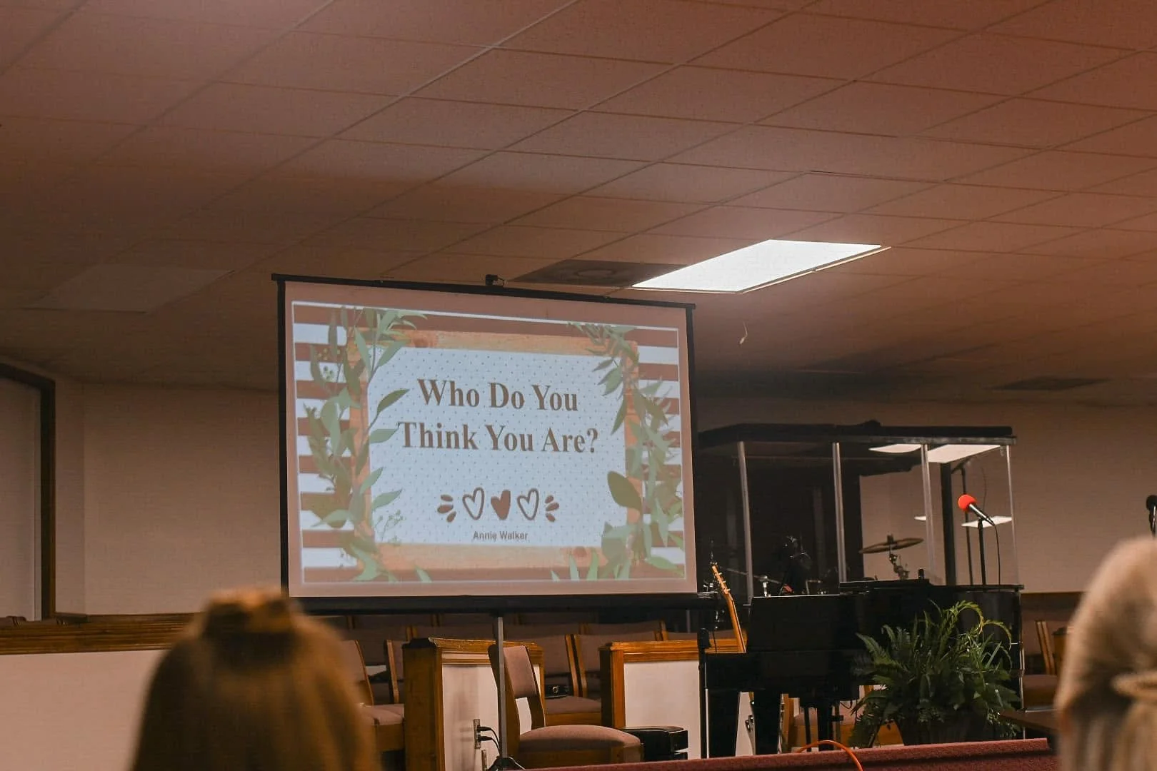 A presentation slide on a large screen with the title "Who Do You Think You Are?" by Annie Walker, with a decorative leafy border, in a room with chairs and musical instruments.