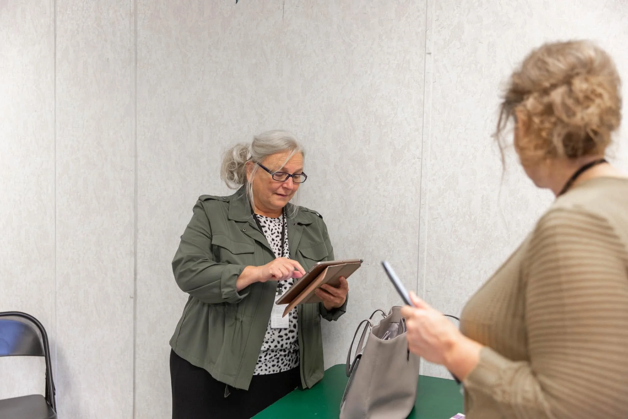 Two women standing at a table, engaged in a conversation, one holding a clipboard and the other with a pen, in a room with plain white walls and empty chairs nearby.