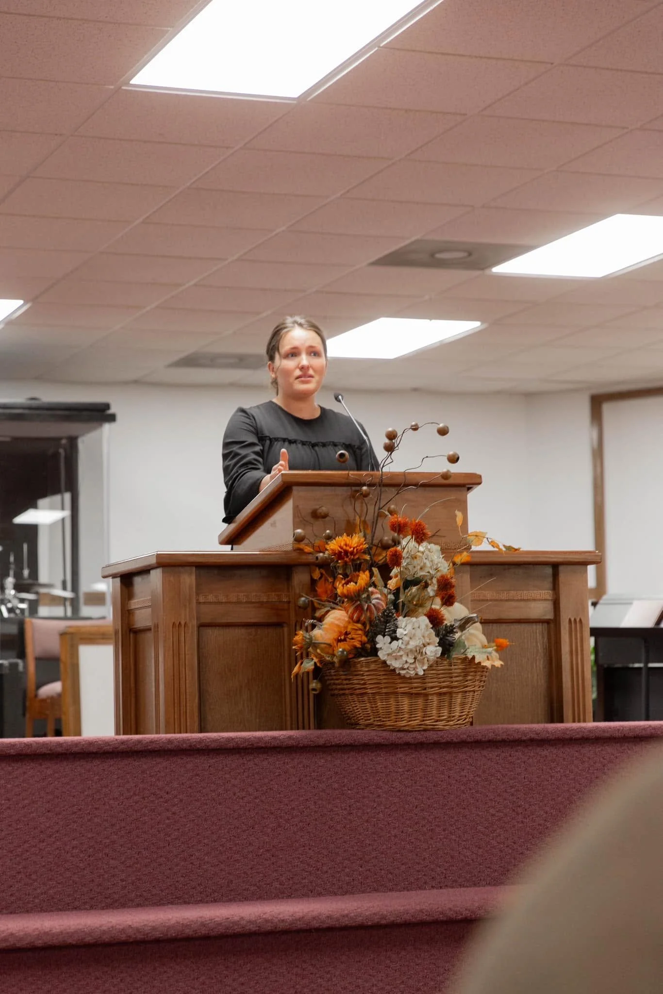 A woman standing behind a wooden podium with a floral arrangement at its front, in a room with a drop ceiling and rectangular fluorescent lights.