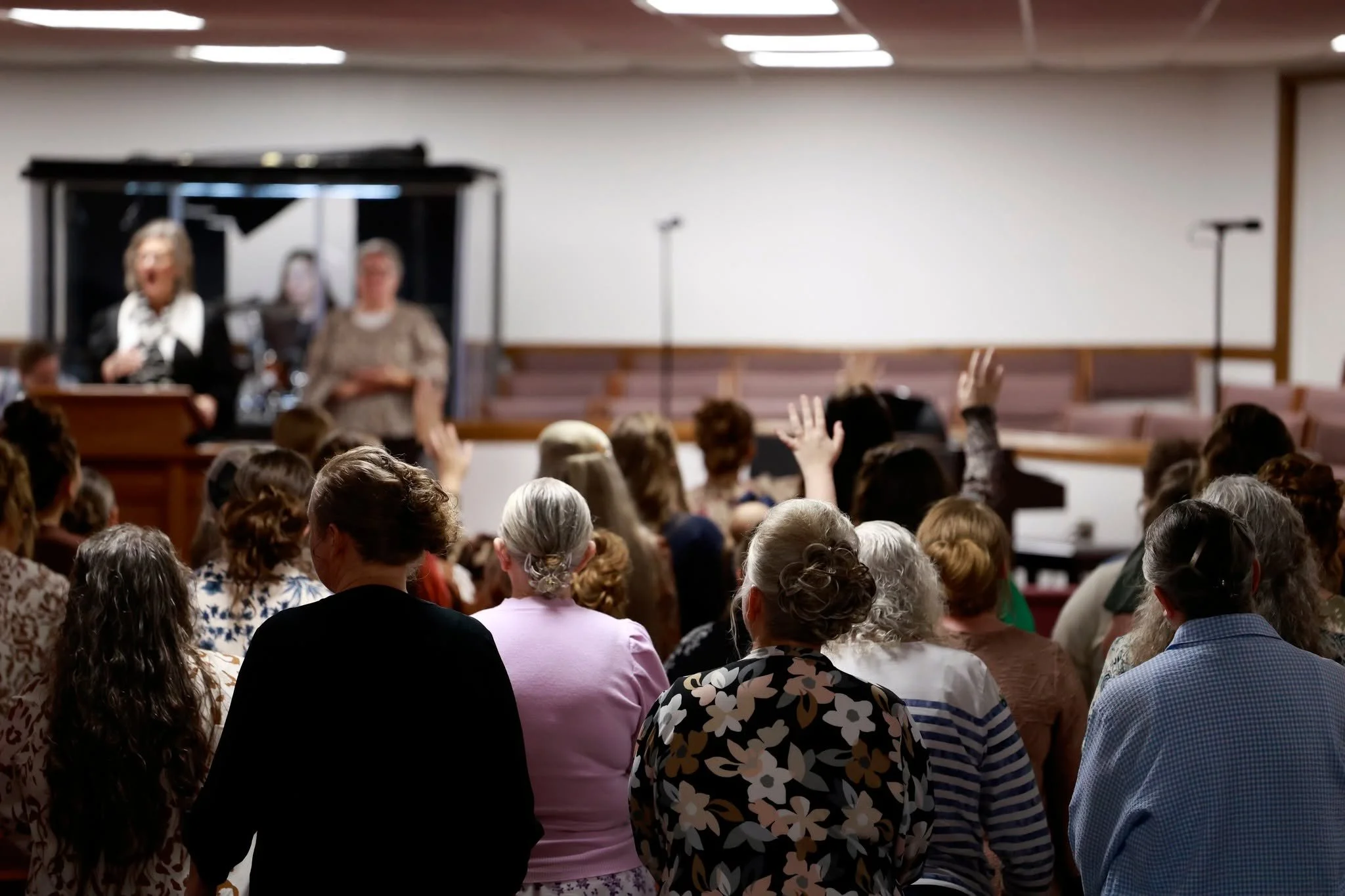 A diverse group of people attending a presentation or lecture in a conference room, with some raising their hands. The speaker is a woman standing at the podium near the stage, with a piano in the background.