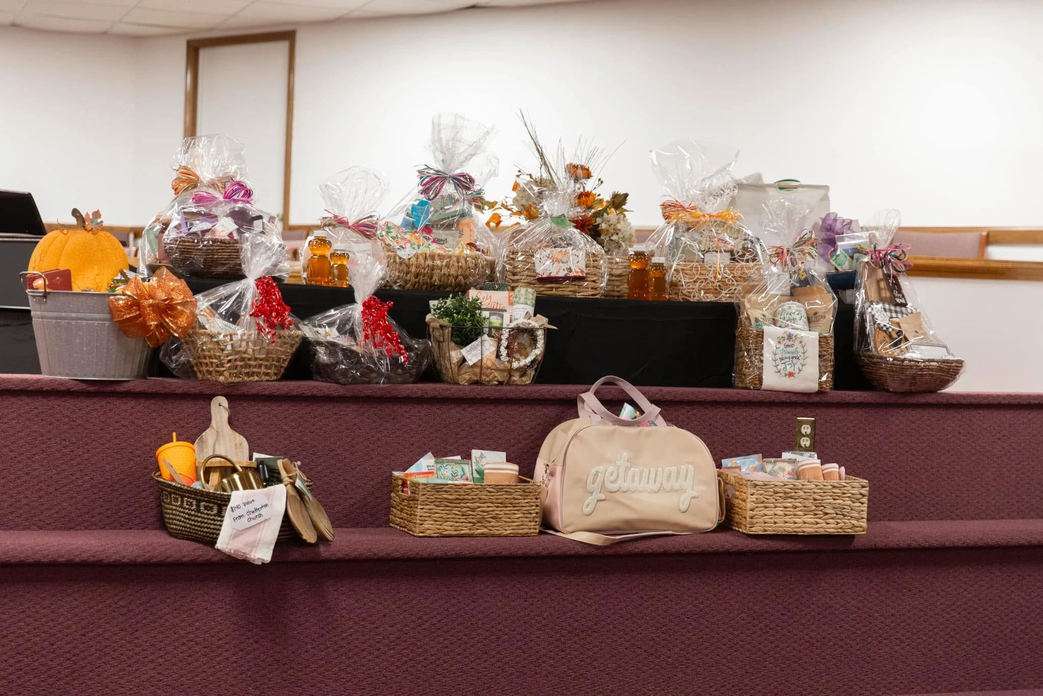 Display of multiple gift baskets and items on tiered steps, including a pumpkin, bows, jars, and holiday decorations, with a beige bag labeled 'getaway' in front.