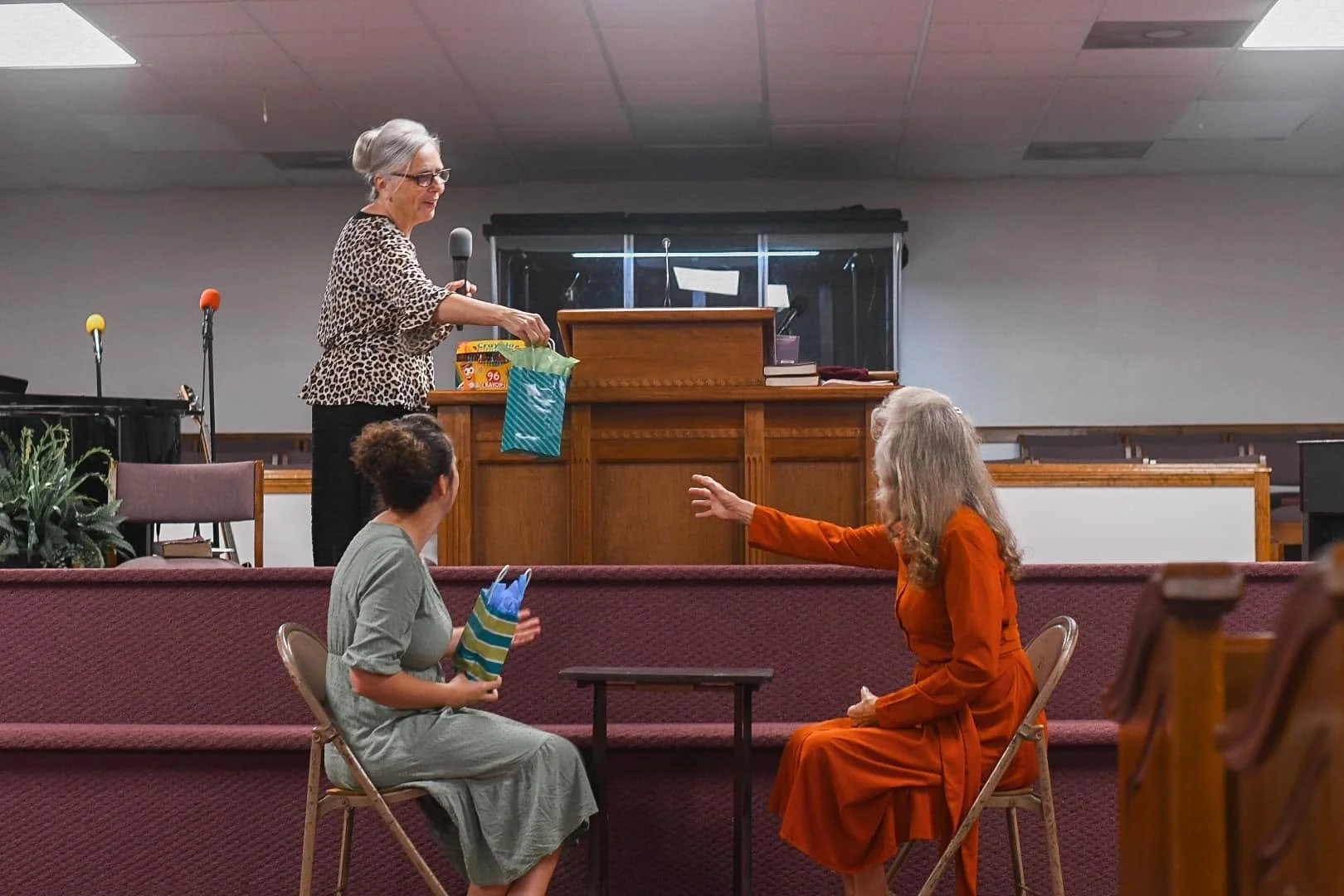 An elderly woman with glasses in leopard print top hands out gifts to two women seated on chairs in a church setting, with a piano and microphones in the background.