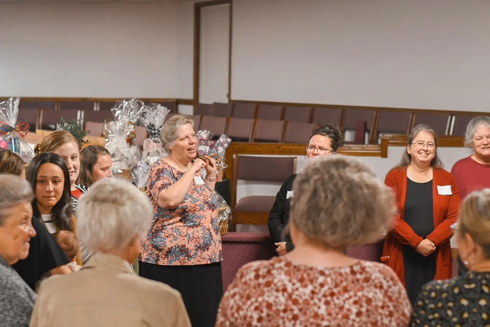 A group of women smiling and standing in a semi-circle at an indoor gathering, with gift baskets on a table behind them.