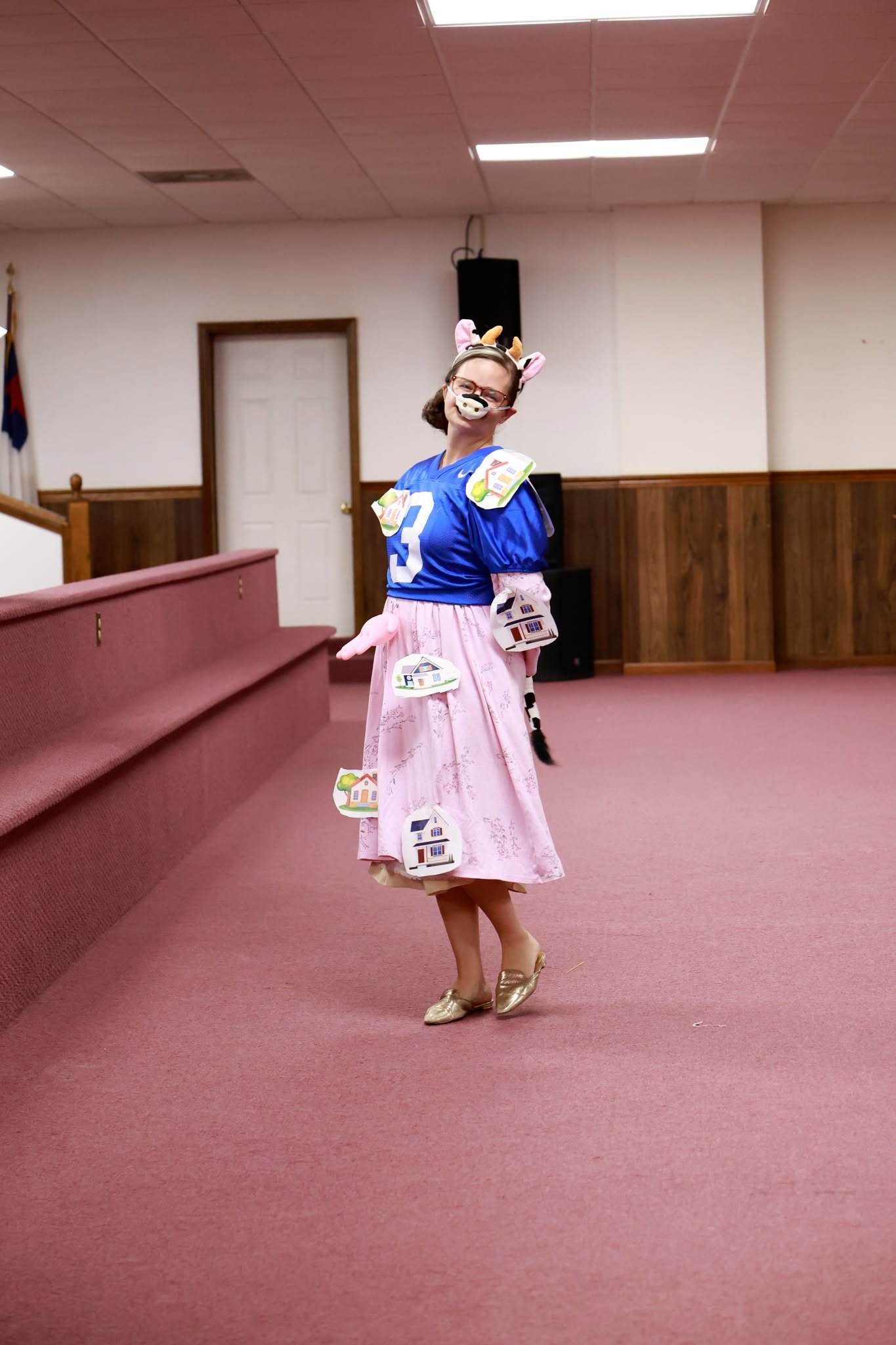 A woman standing in a room decorated with pink and white, wearing a blue football jersey with the number 3, a pink skirt with house emblems attached, a cow and deer headband, a mask resembling a cow's nose, and gold shoes. She is smiling and posing w