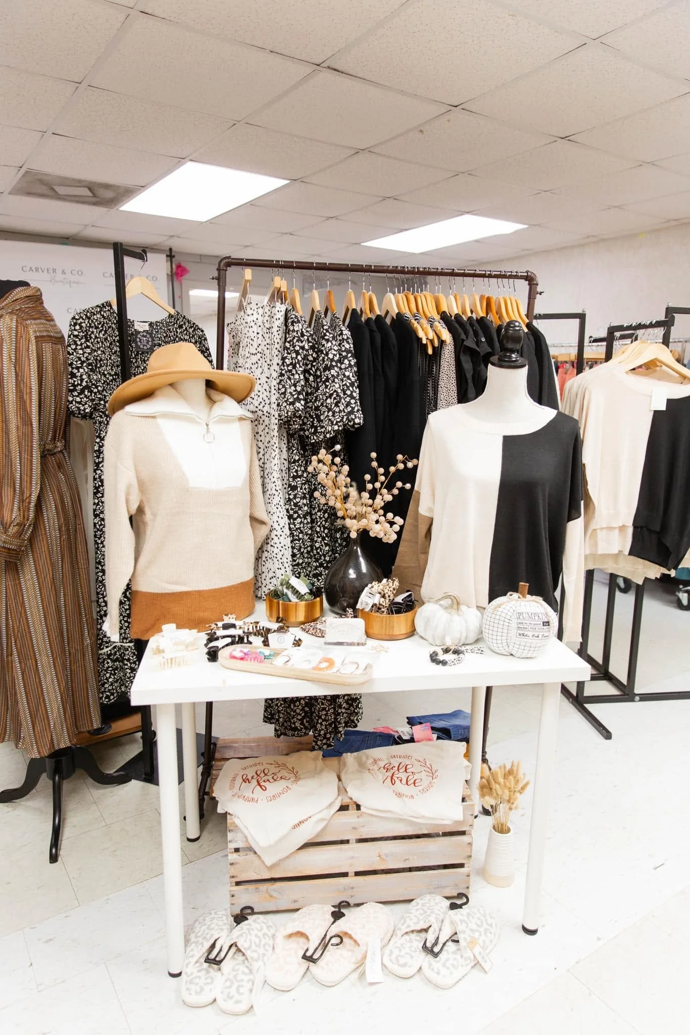 Display of women's clothing, accessories, and home decor items in a retail store. Bottles, flowers, and decorative signs are on the table in the foreground. Clothing racks with black and patterned dresses are in the background.