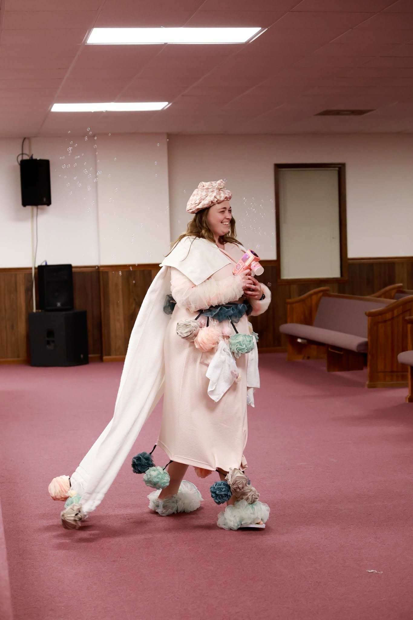A woman in a pink hat and a cream-colored dress is holding a bubble gun and smiling. She is dressed in a whimsical costume decorated with fabric flowers on her shoes, dress, and cape. The setting is a room with wood-paneled walls, benches, and speake