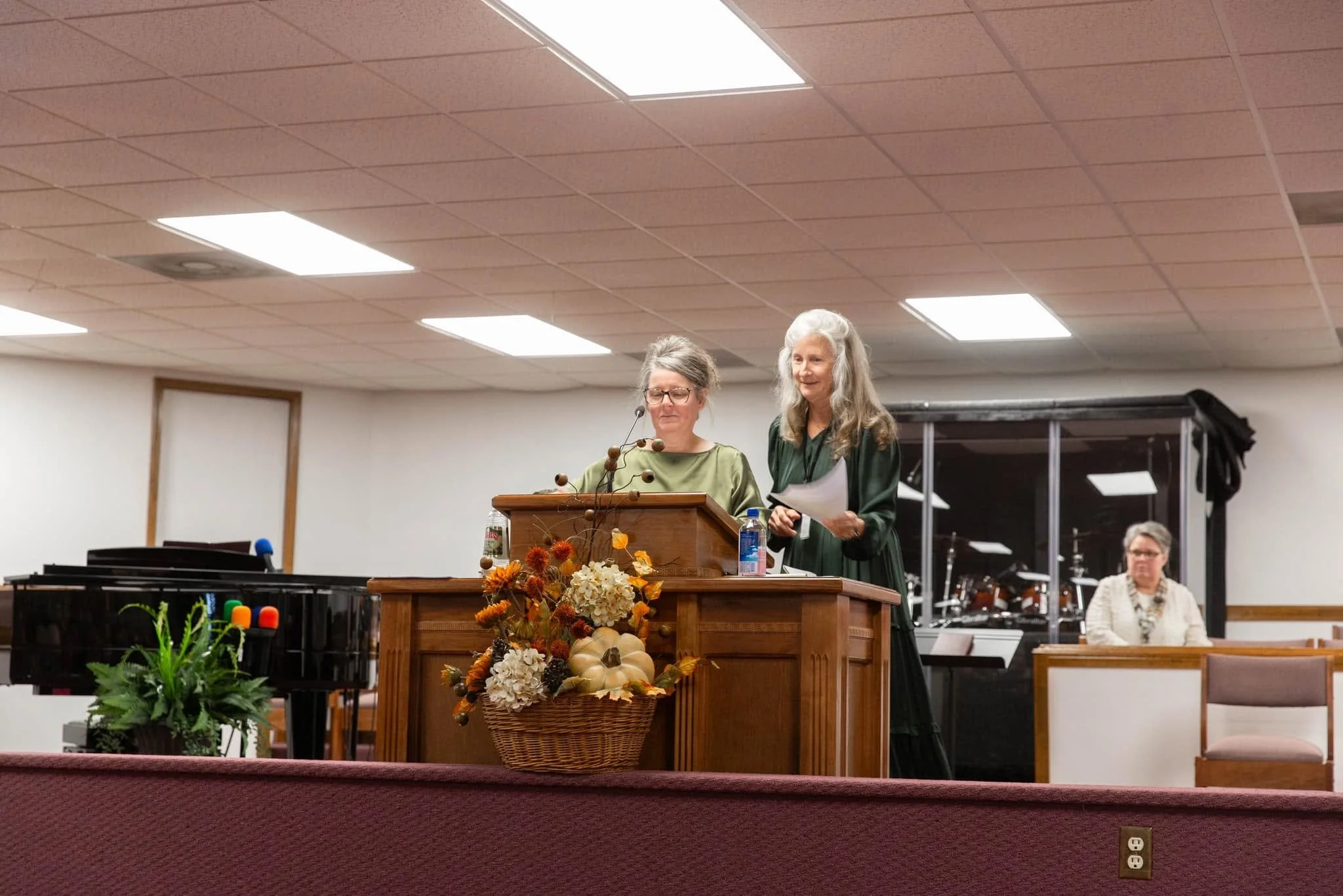 Two women standing behind a wooden podium with flowers, one reading from papers, in a room with people and musical instruments in the background.