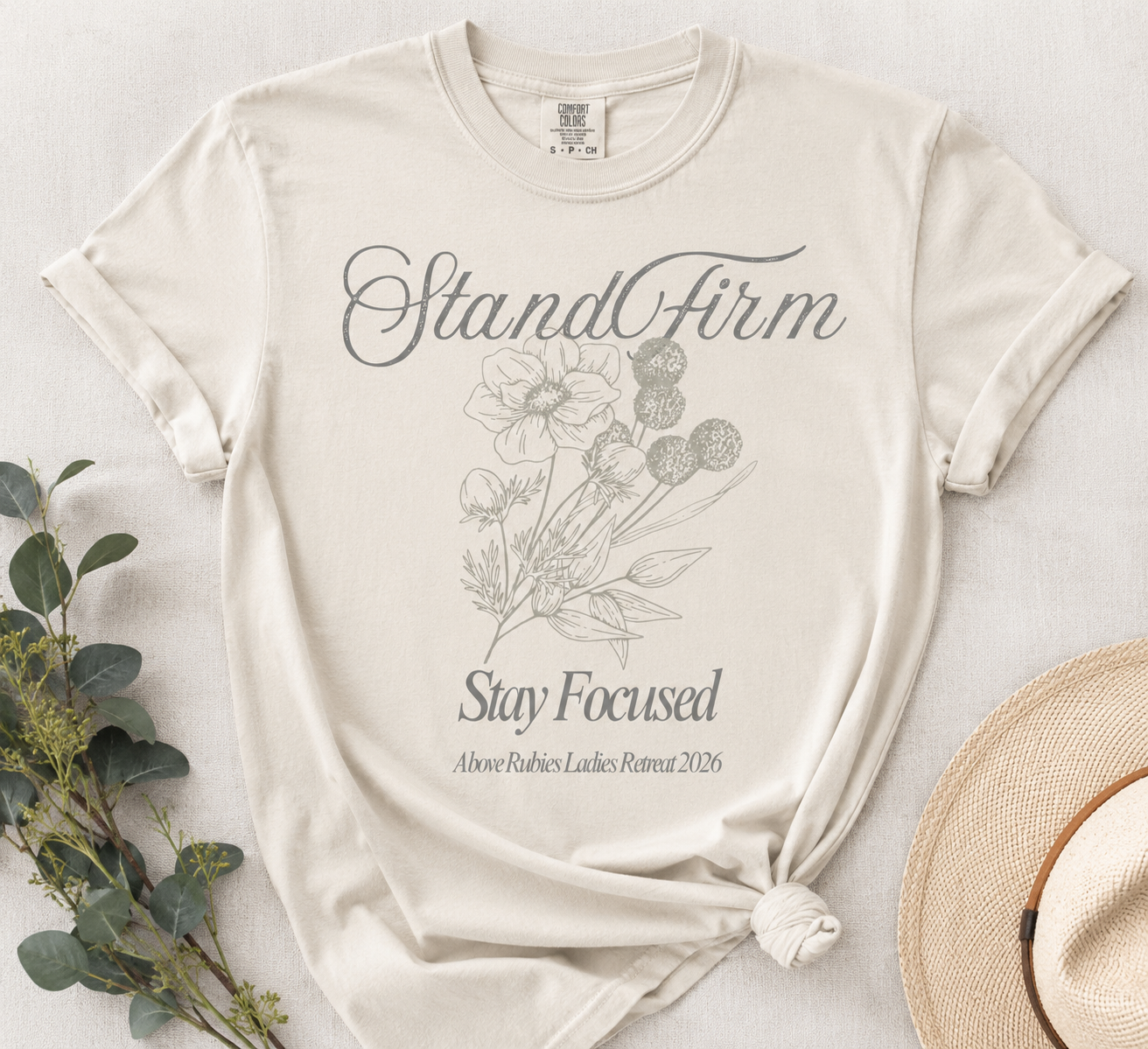 A beige T-shirt with the words 'Stand Firm' and 'Stay Focused,' a floral illustration, and the text 'Above Rubies Ladies Retreat 2026' printed on it, laid flat alongside a straw hat and some greenery.