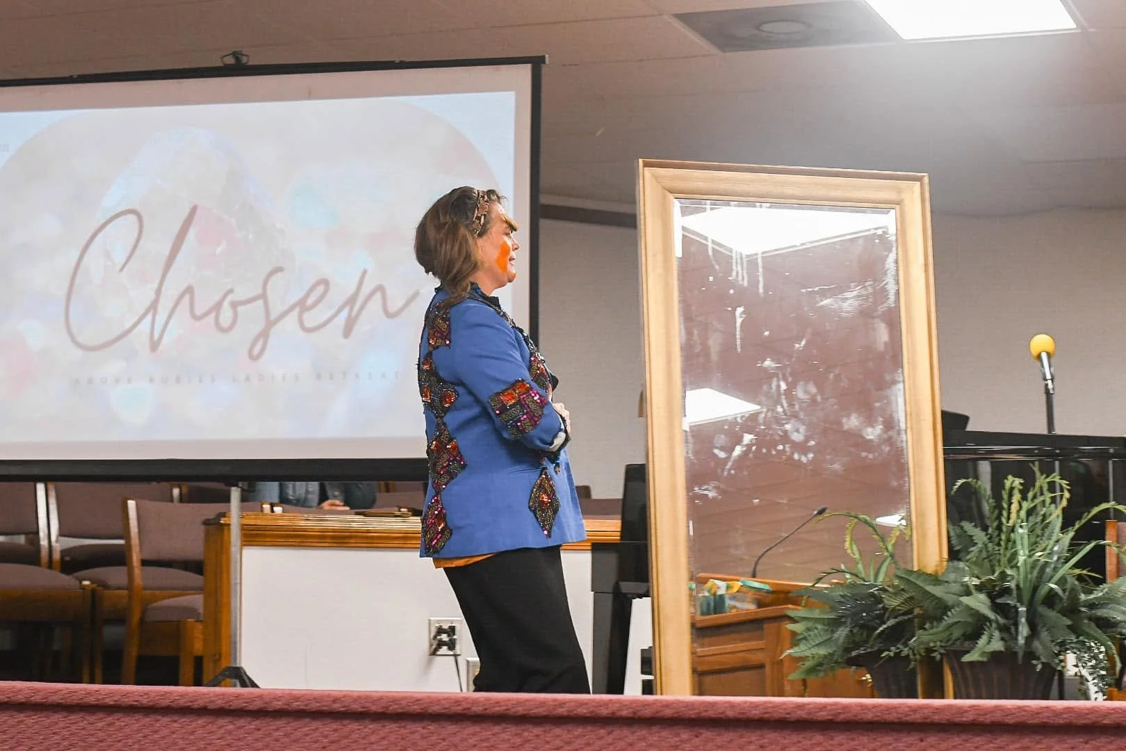 A woman standing on a stage in front of a large projection screen with the word "Chosen" on it, next to a large mirror, with a potted plant nearby.