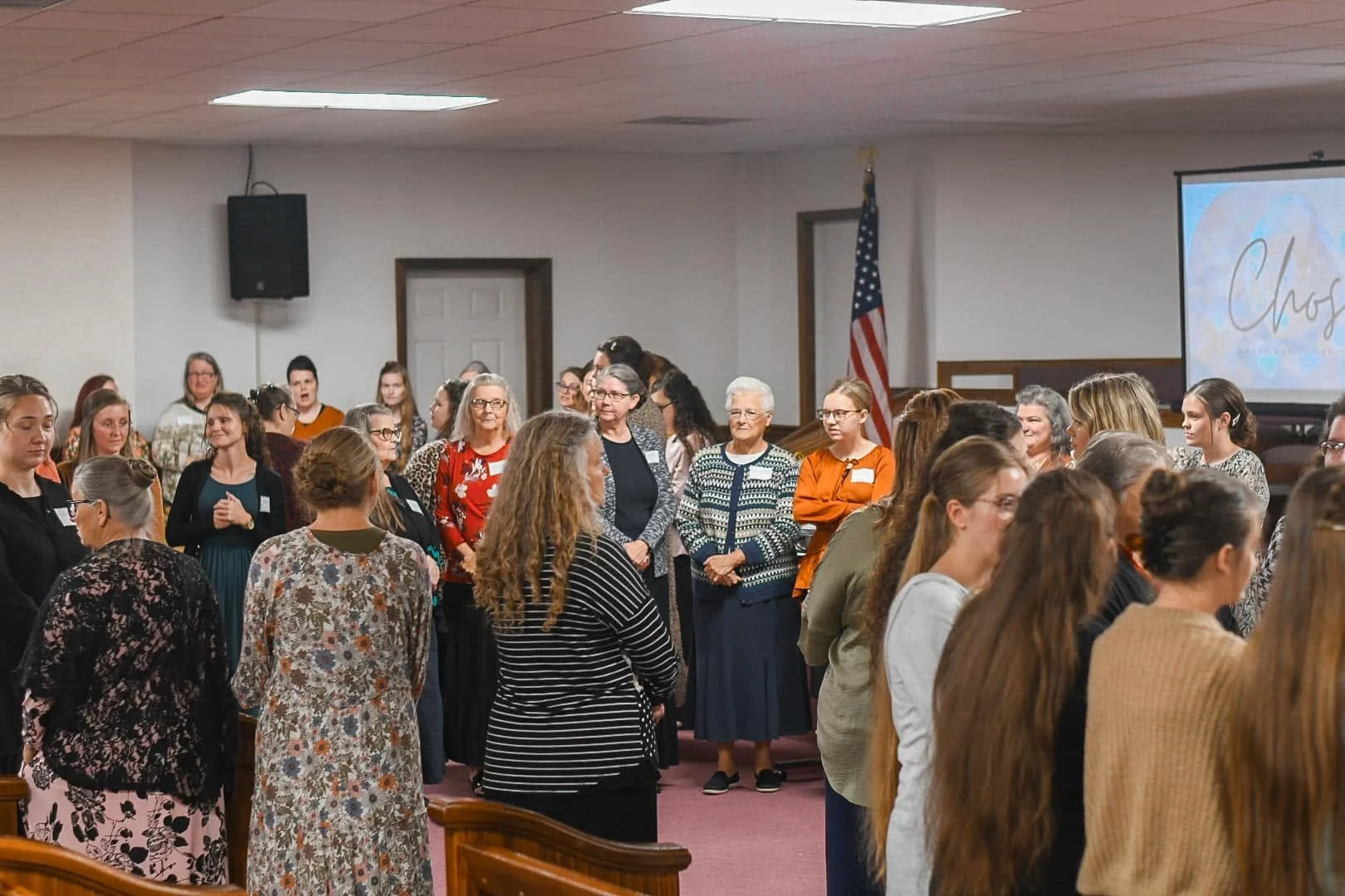 A large group of women gathered in a church or conference room, standing in prayer or reflection. The room has a projector screen displaying the word 'Chog' with a background of clouds and a sunset, and an American flag is visible in the background.