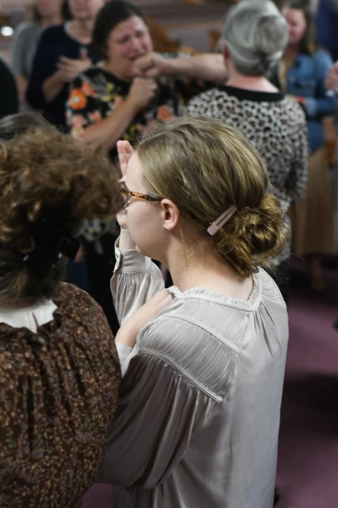Women in a religious or cultural ceremony, with one touching or embracing another, in a crowded indoor setting.