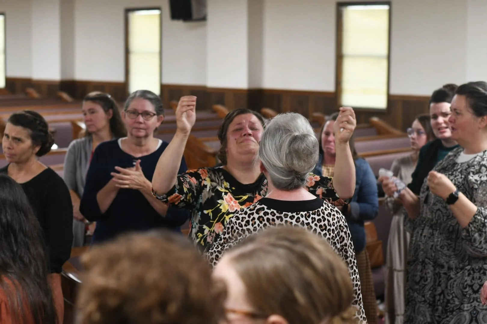 A woman is crying and raising her hands in front of a group of women in a church or community hall, with some holding tissues or prayer books.