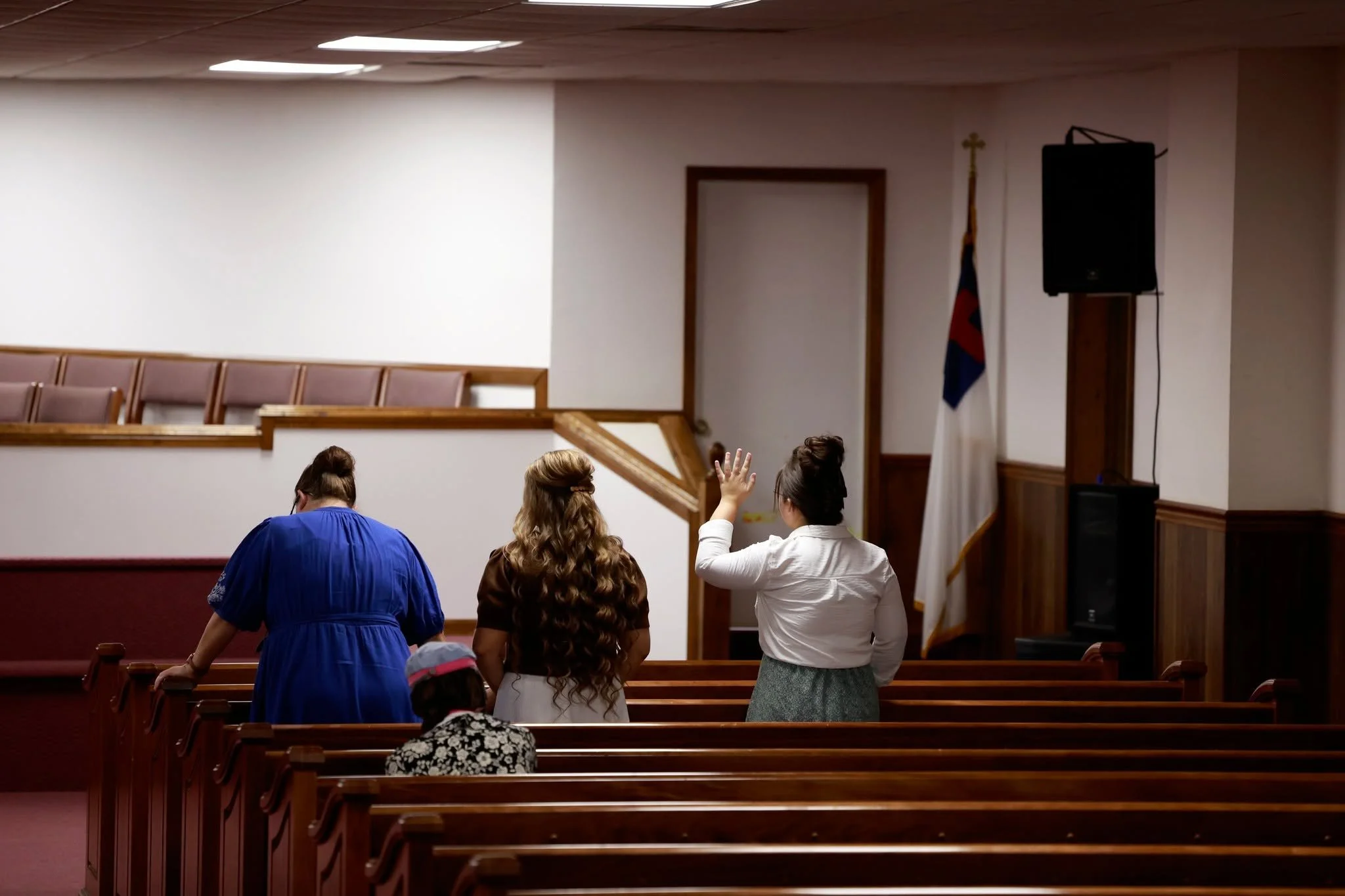 People standing and sitting inside a church, with some raising hands, near a wall with a flag and a speaker.