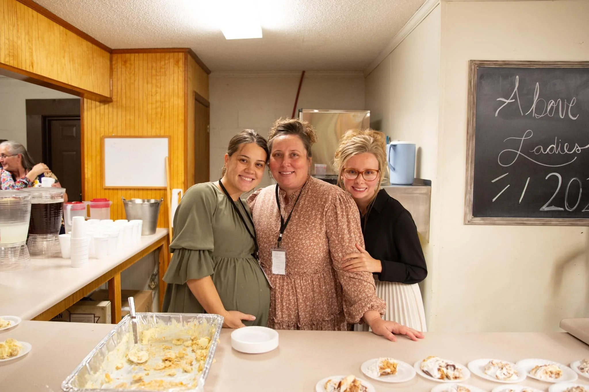 Three women standing together in a kitchen, smiling at the camera. One woman is wearing glasses, and the other two are standing close to her with their arms around her. In the background, a woman with gray hair is sitting at a table, and there are fo