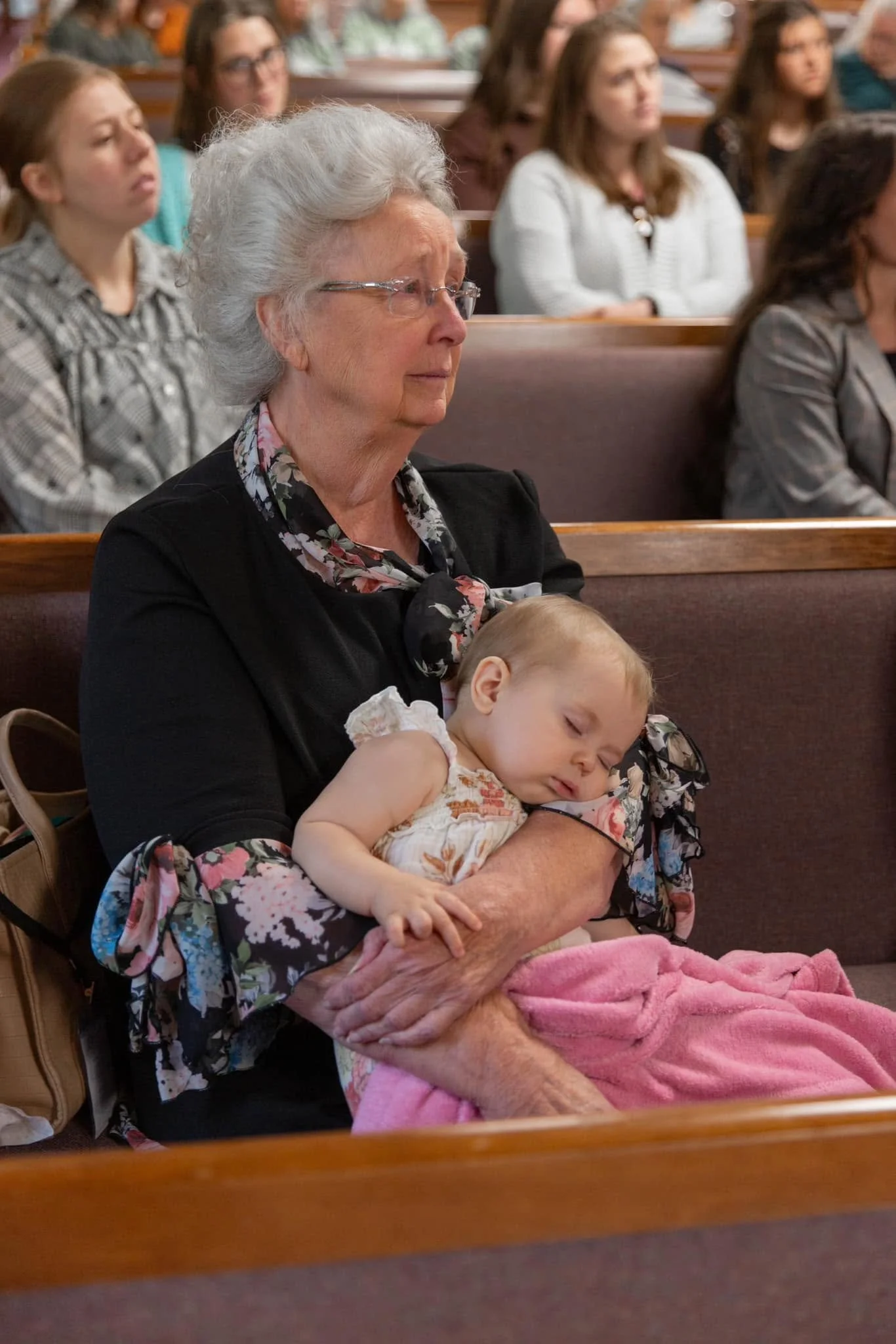 An elderly woman with gray hair and glasses, wearing a black jacket and floral blouse, is sitting in a church pew holding a sleeping baby girl wrapped in a pink blanket.