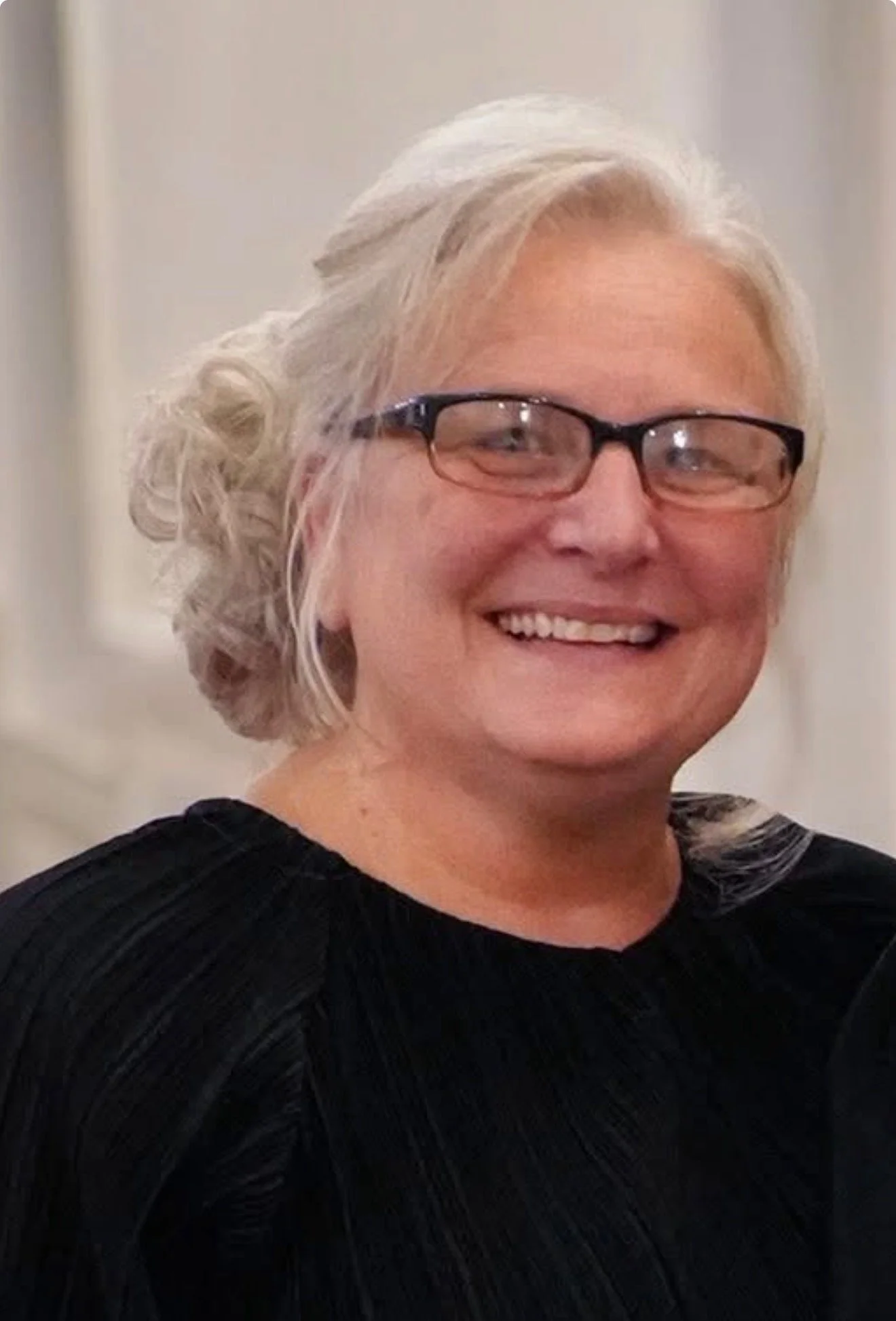 A smiling woman with gray hair styled in soft curls, wearing black-rimmed glasses, and dressed in a black top.