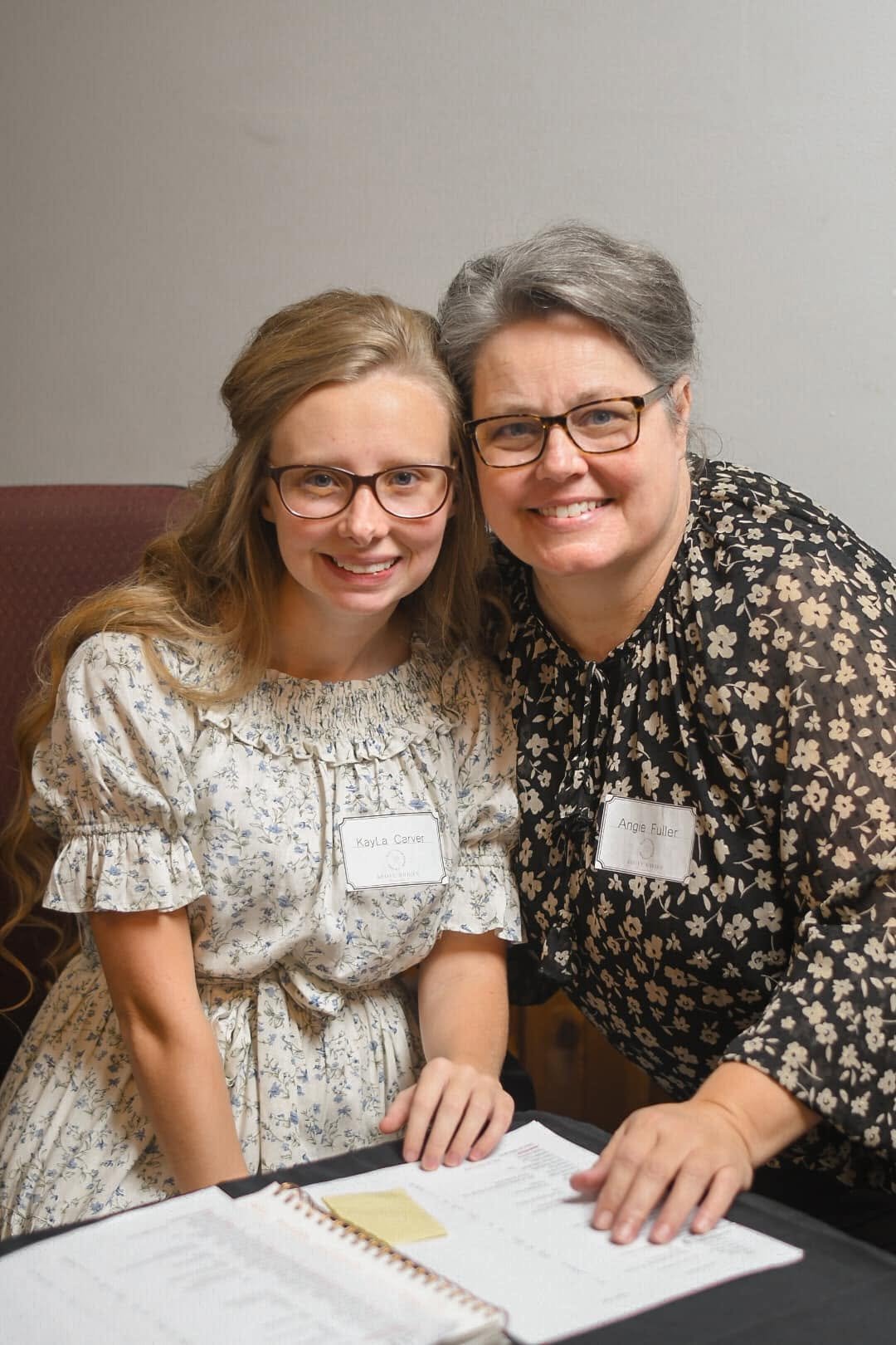 Two women, Kayla Carver and Angie Fuller, smiling and sitting at a table with documents, wearing glasses and floral dresses, in a room with plain walls.