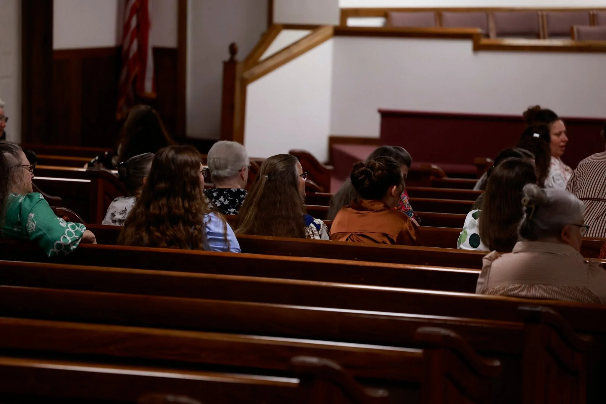 People sitting in pews inside a church or auditorium, listening attentively, with wooden railings and an American flag in the background.