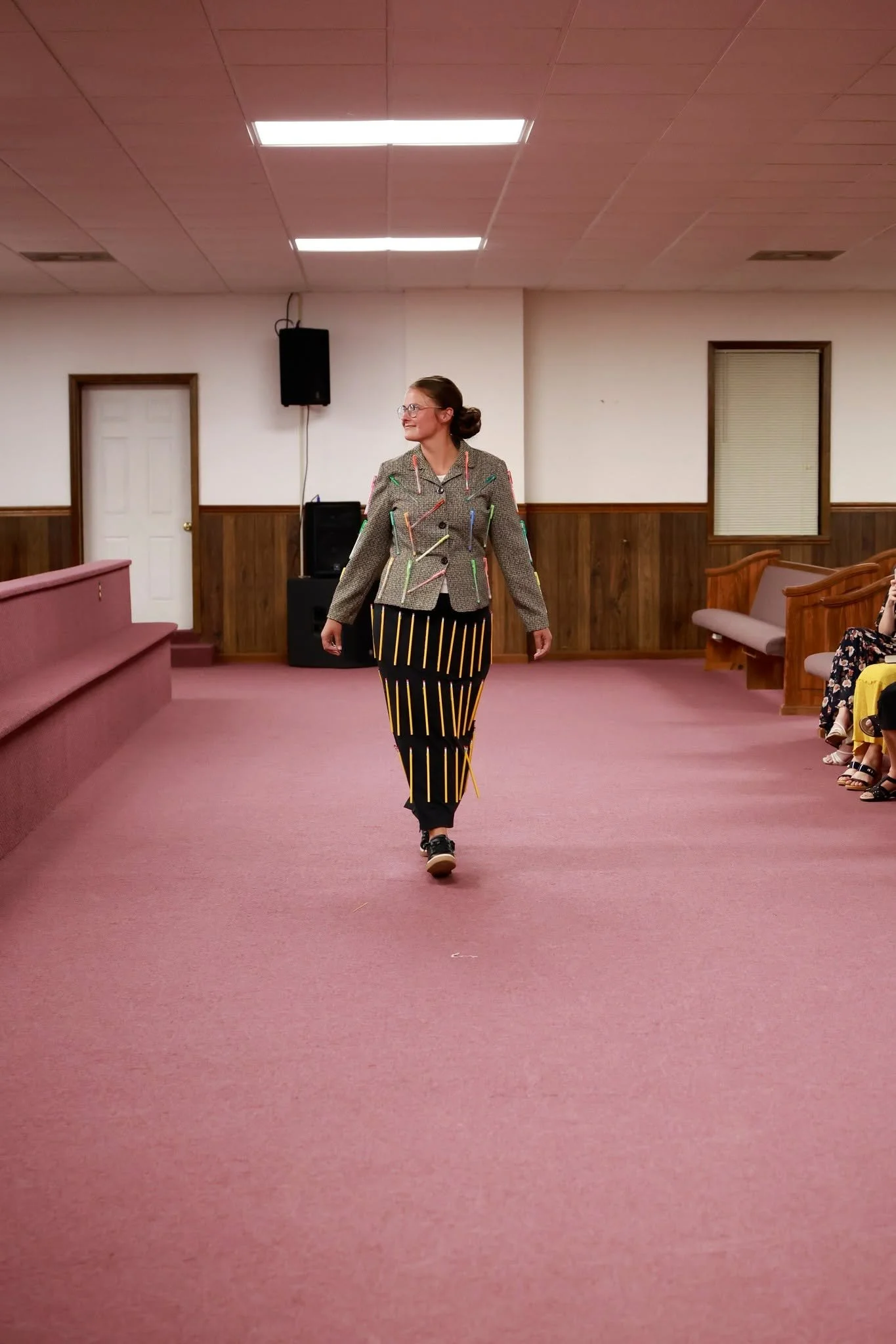 Woman walking down an aisle in a room with wood-paneled walls, pink carpet, and fluorescent ceiling lights, wearing a blazer adorned with colorful pens and black pants with yellow stripes.
