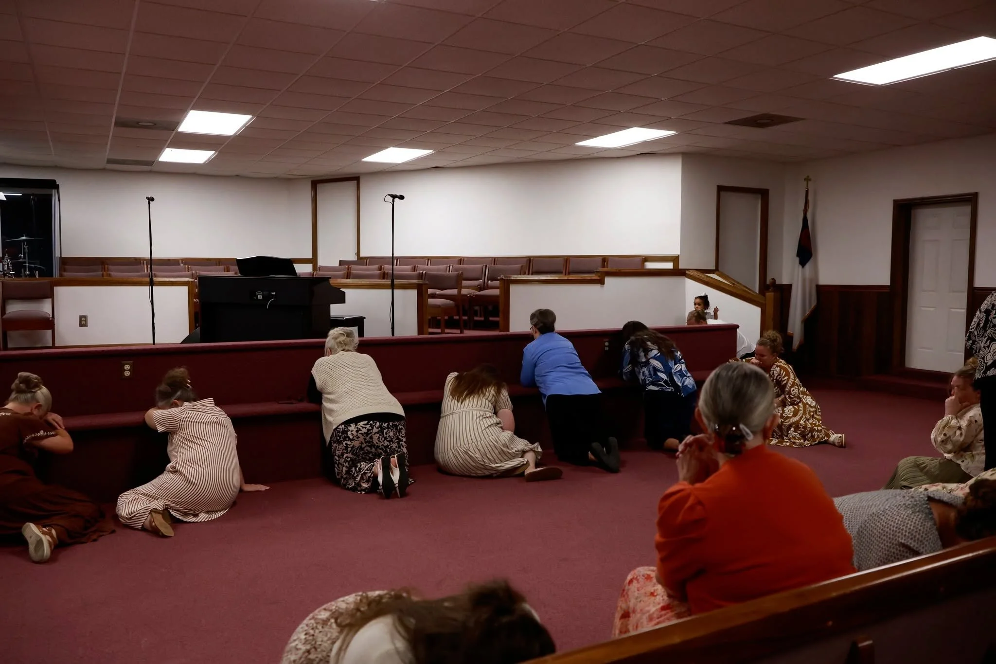 People kneeling and sitting on the floor in a church or auditorium. The stage has a piano, microphones, and a drum set. The room has a drop ceiling with fluorescent lights and evangelical flags.