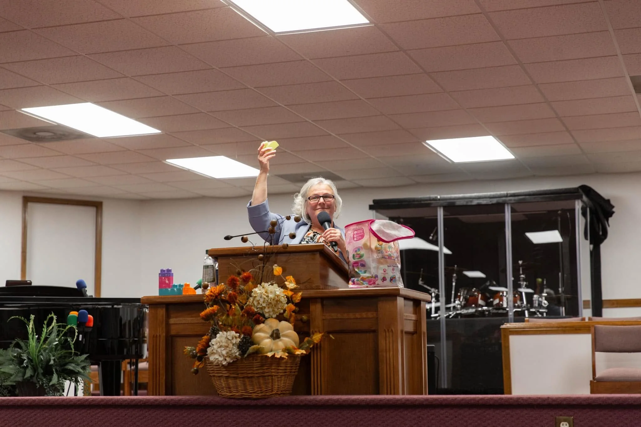 A woman with glasses and gray hair speaking at a podium decorated with flowers and fall decorations, holding a microphone and a yellow object, in a room with a stage, musical instruments, and a black roofed shelf in the background.