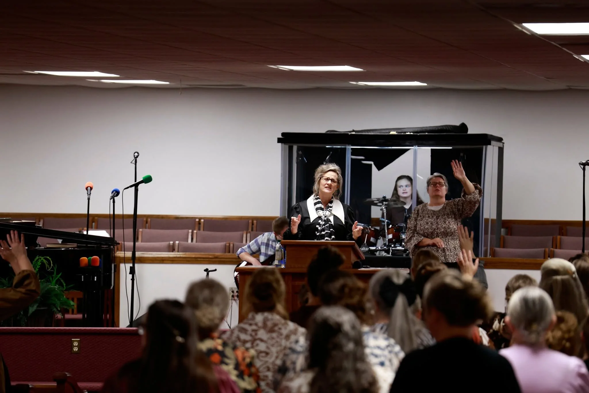 A church service or religious gathering with women speaking or praying at the front, surrounded by an audience of children and adults, with microphones and musical instruments nearby.