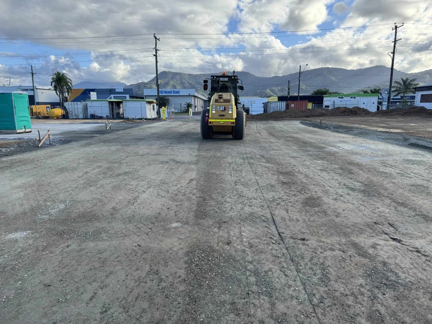 A construction site with a Caterpillar compactor in the center, dirt and gravel ground, temporary structures, and utility poles under a partly cloudy sky with mountains in the background.