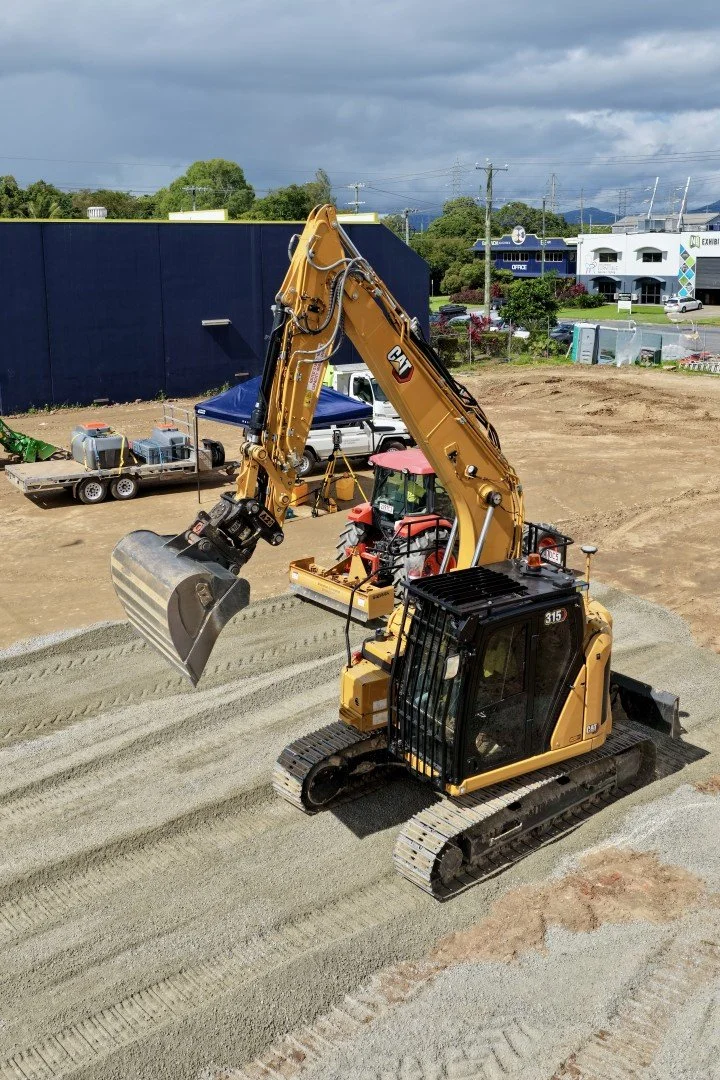 A yellow Caterpillar excavator with a tracked base and a hydraulic arm, working on a construction site with gravel ground and a scoop attached to the arm.