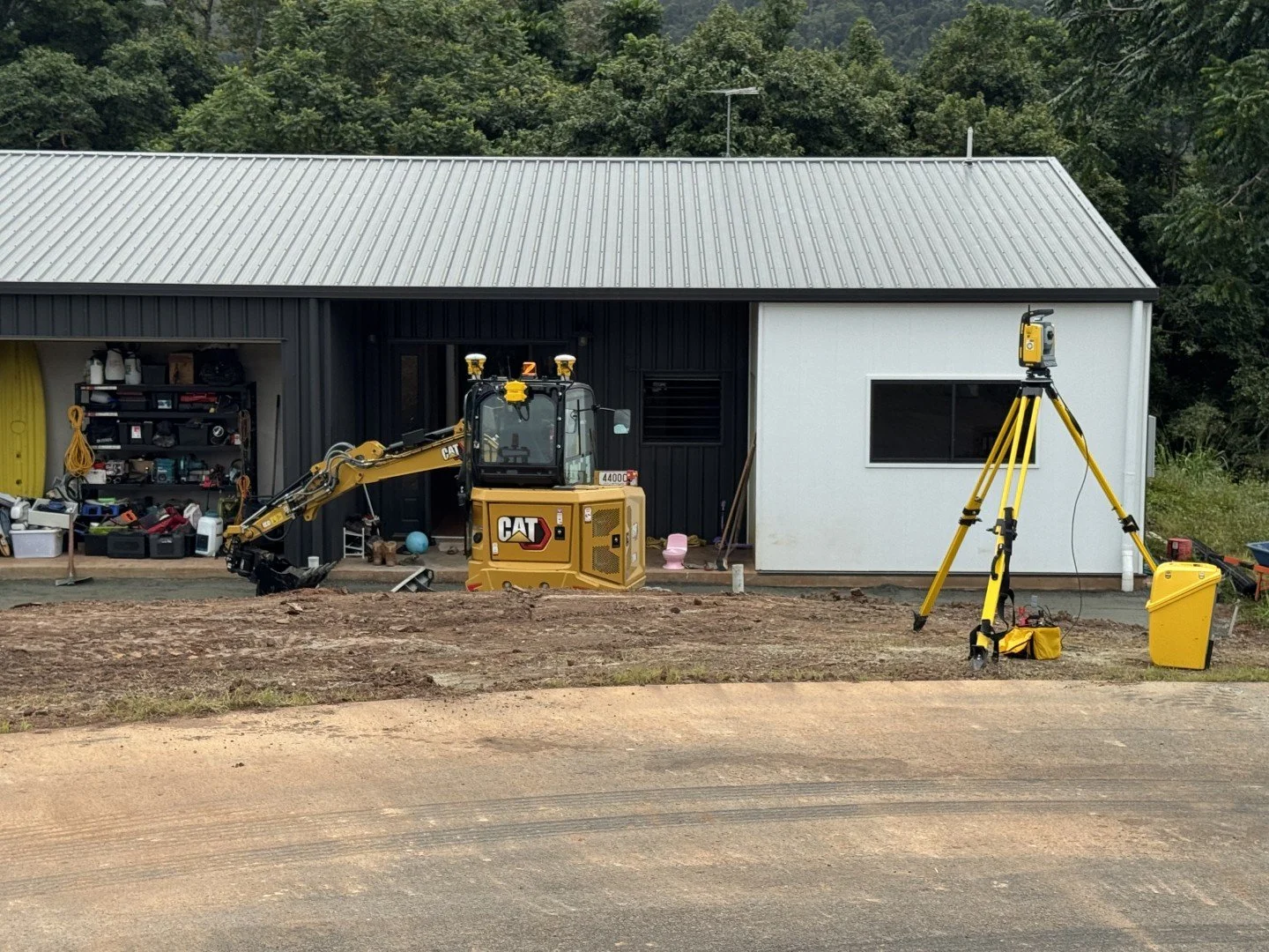 Construction site in front of a modern house with black and white exterior walls. A small excavator is parked near a wall shelves filled with tools, and a surveying tripod is set up on the ground. There is dirt and gravel on the ground, indicating ongoing construction work, with a wooded area in the background.