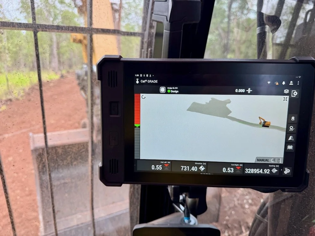 Close-up of a tablet screen inside a construction or logging vehicle, displaying a digital map with an excavator moving along a curved path on a map, with trees and forest outside in the background.