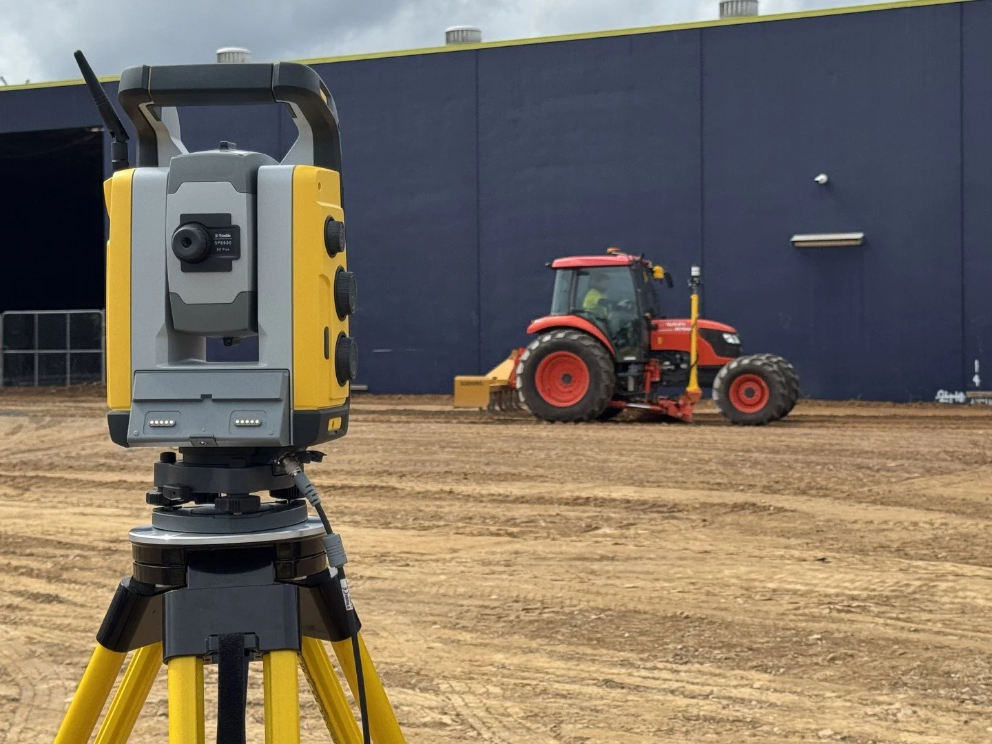 A laser scanning device mounted on a yellow tripod is set up on dirt ground at a construction site. In the background, a red tractor is leveling the soil in front of a blue building.
