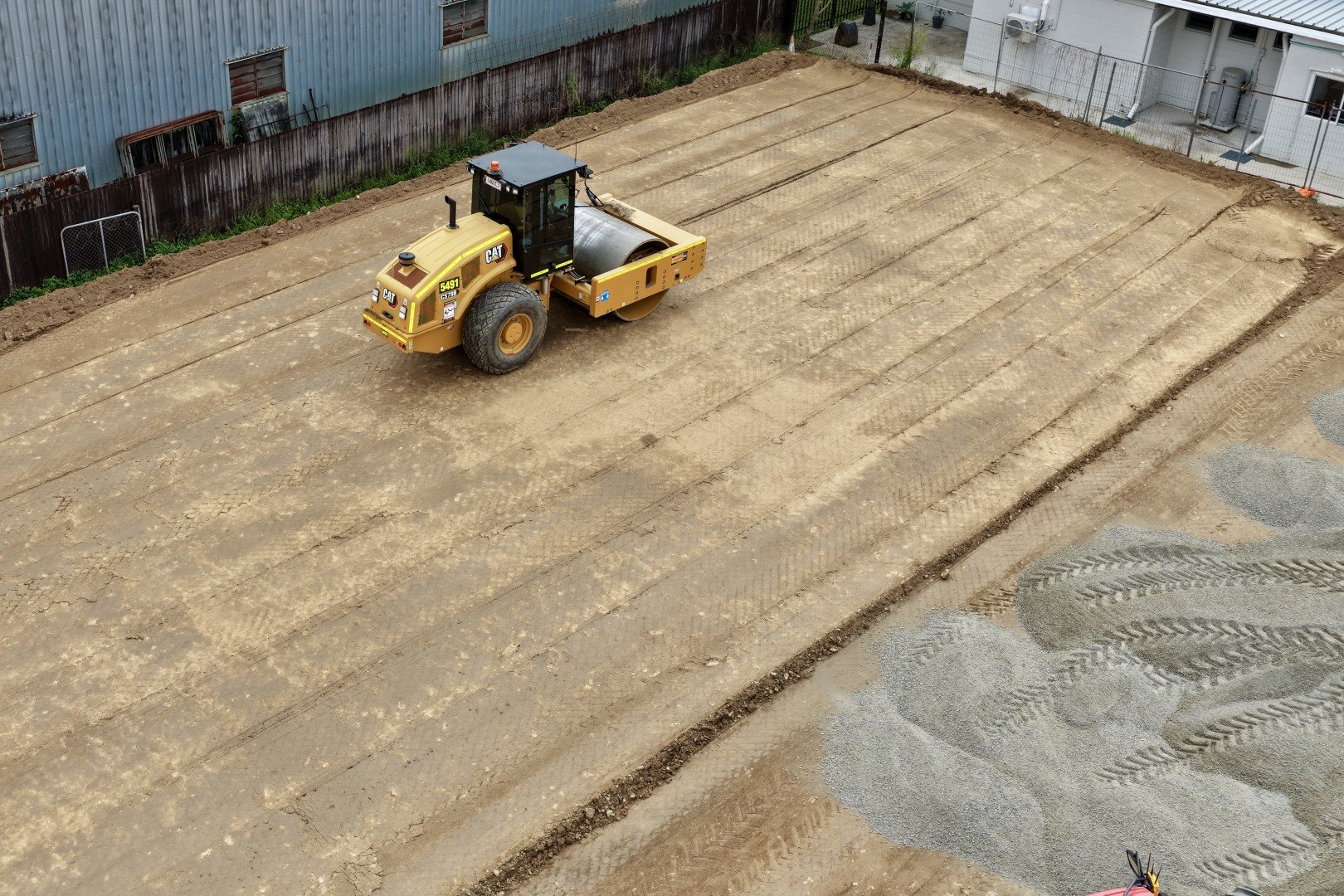 A construction site with a large yellow asphalt roller compactor smoothing out dirt. There are tire marks in the dirt and a chain-link fence in the background.