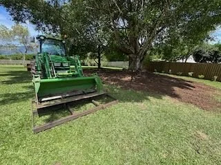 Green lawn mower on grass next to a large tree and a wooden fence.