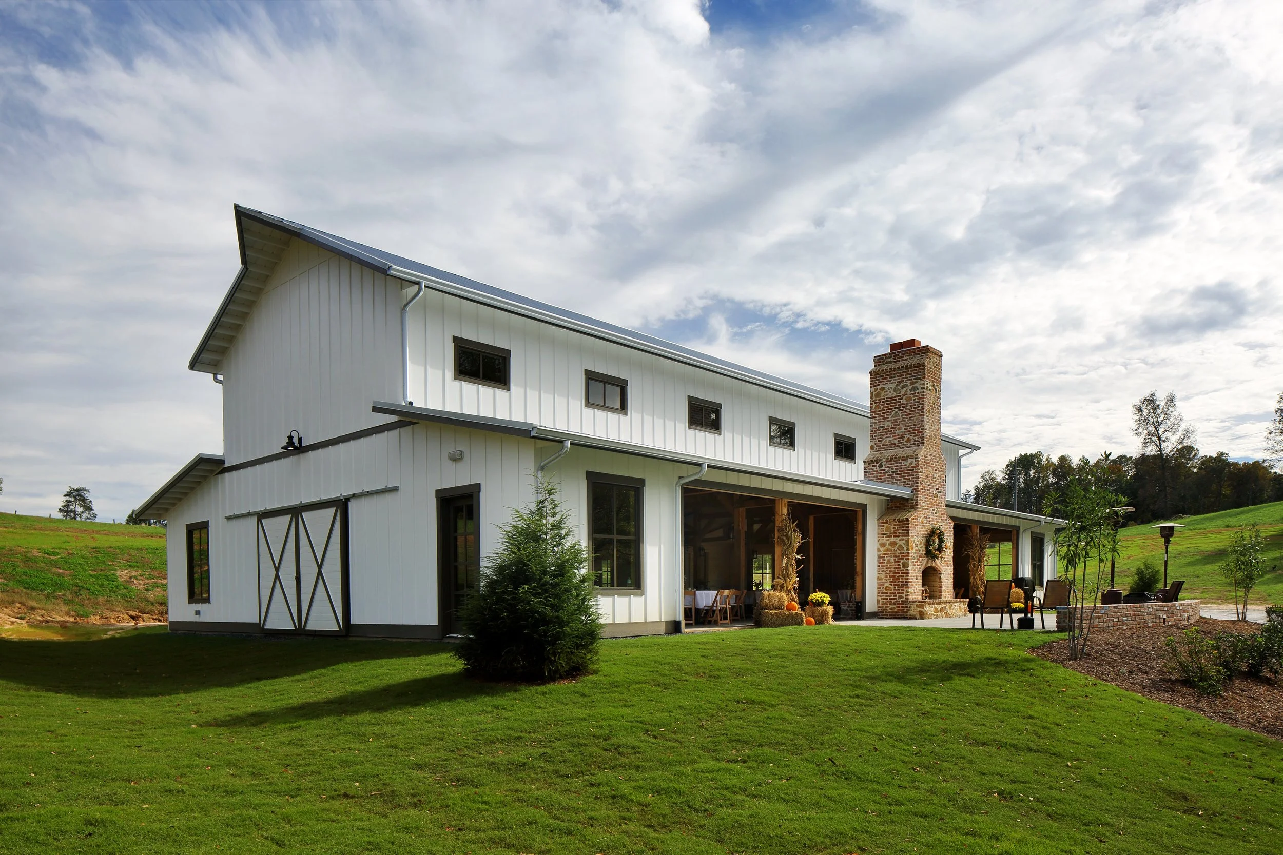 A white modern house with a steep metal roof, large windows, a brick chimney, a patio with chairs and pumpkins, on a green lawn with trees and cloudy sky in the background.